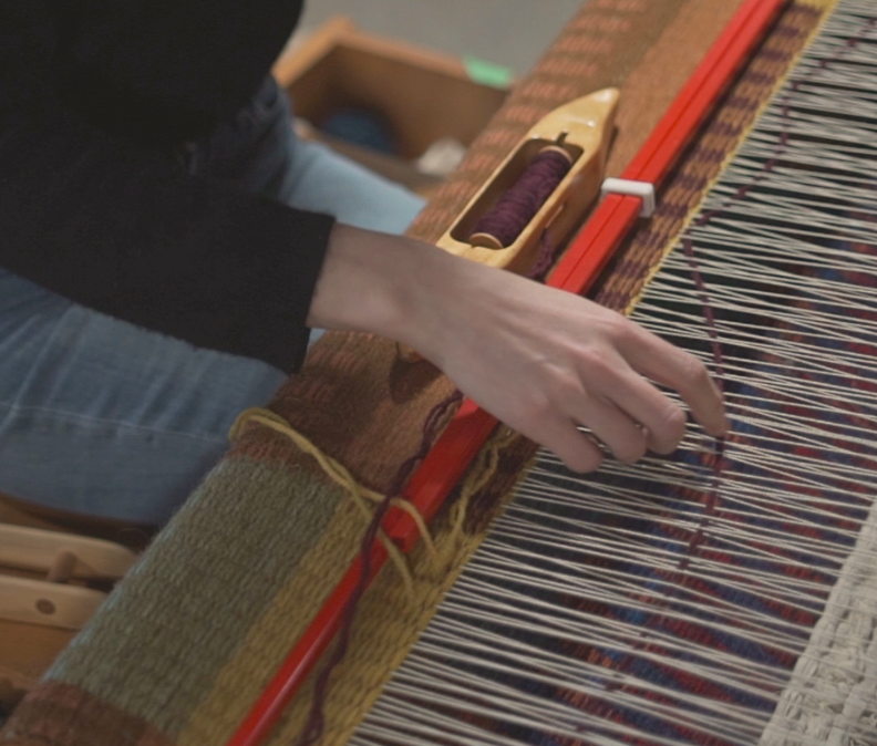 Close-up of person weaving on a loom with colorful woven fabric, hand guiding the warping threads.