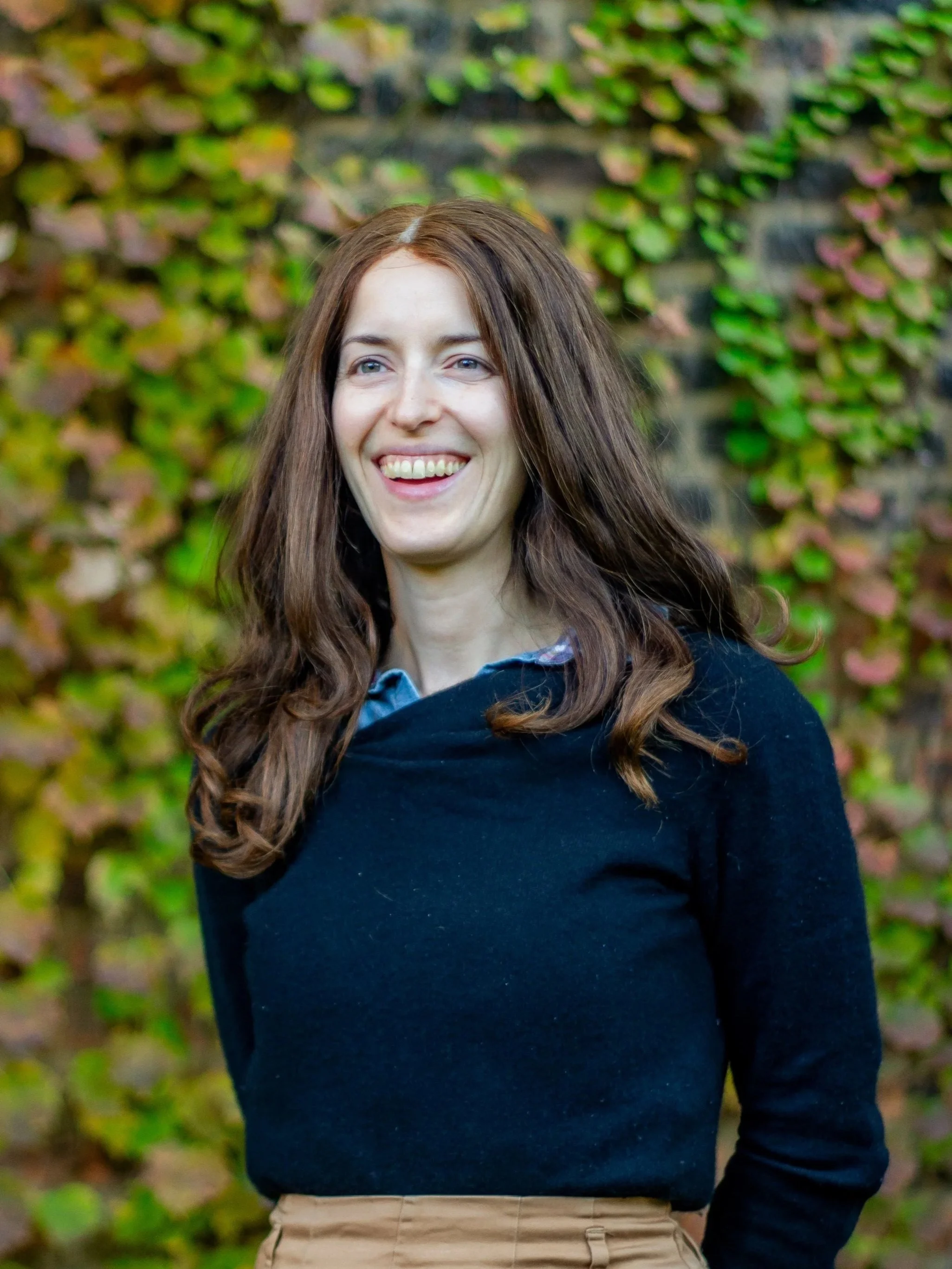 A woman with long brown hair and a bright smile standing outdoors in front of a wall covered with green and purple ivy.