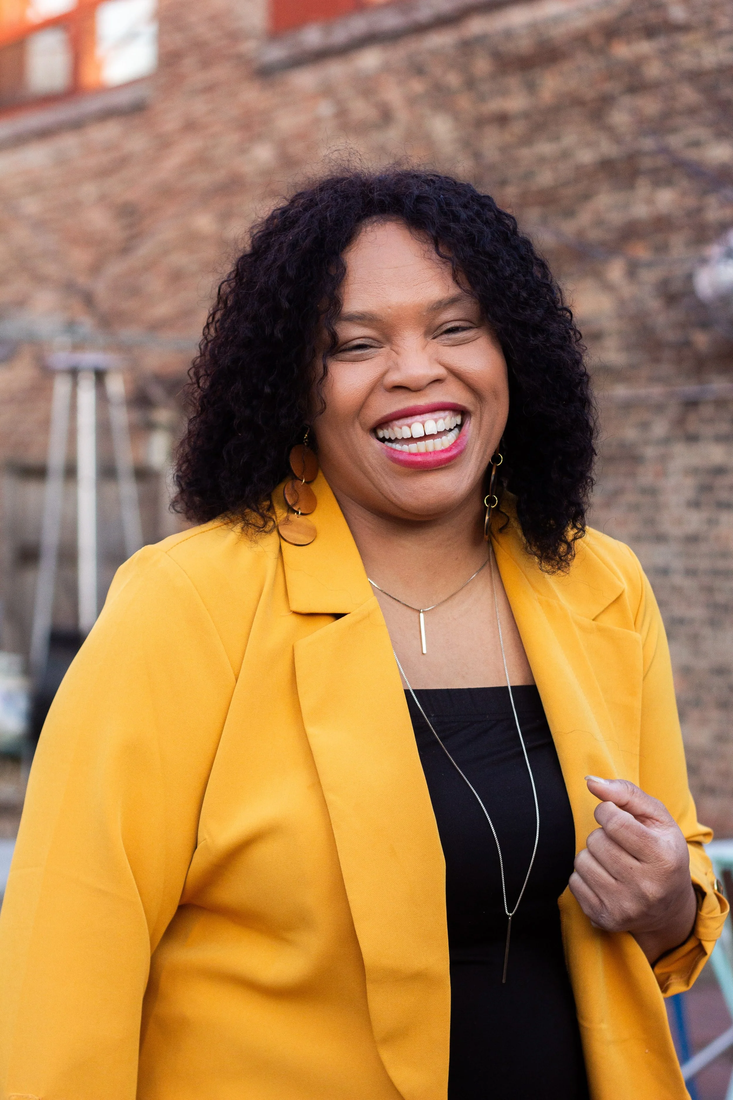 A woman with dark curly hair, wearing a yellow blazer and black top, smiling outdoors with a brick wall in the background.