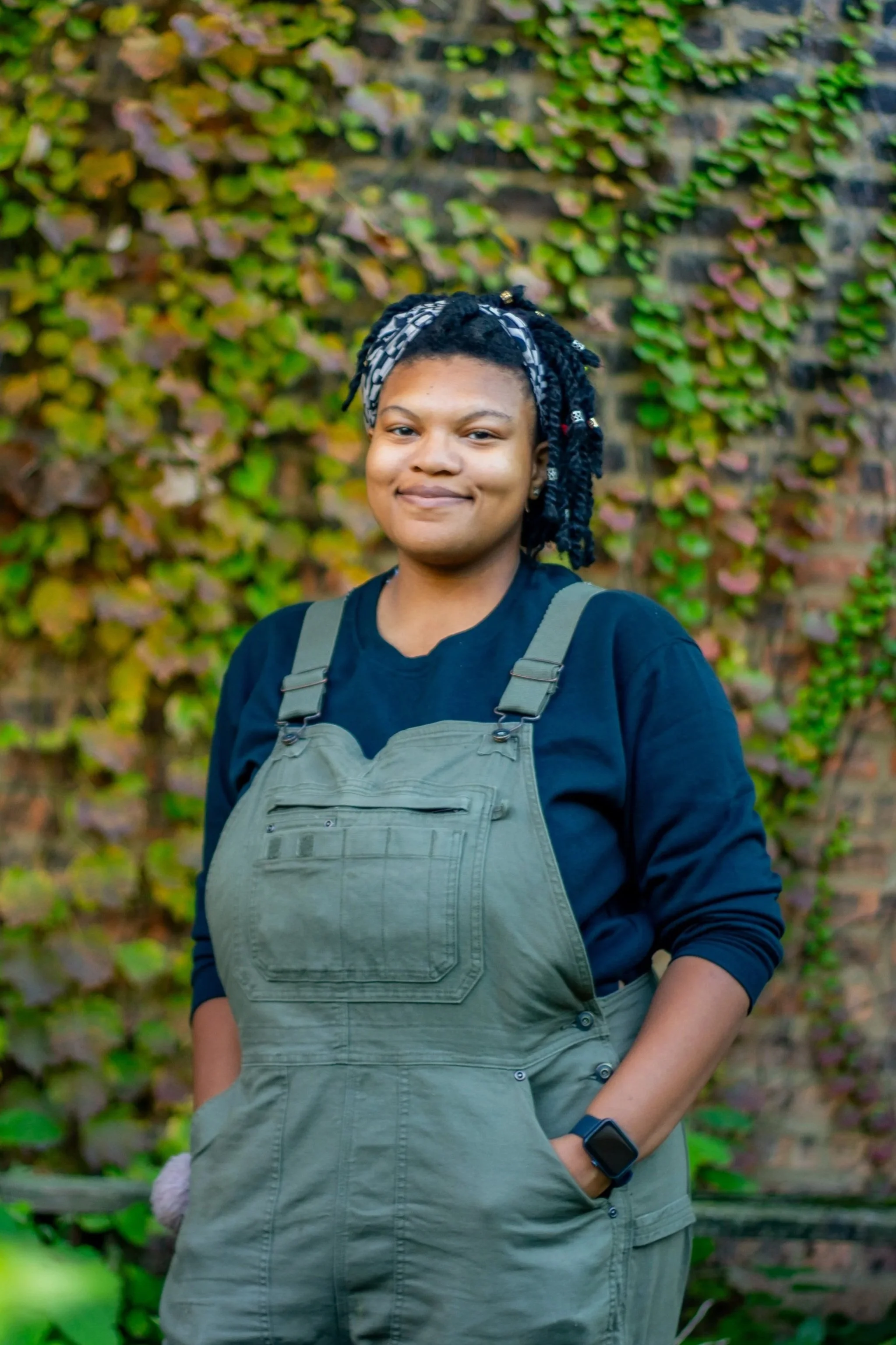 A woman with short, styled hair wearing a black shirt, olive green overalls, and a black smartwatch, standing outdoors against a background of a brick wall covered in green and purple ivy.