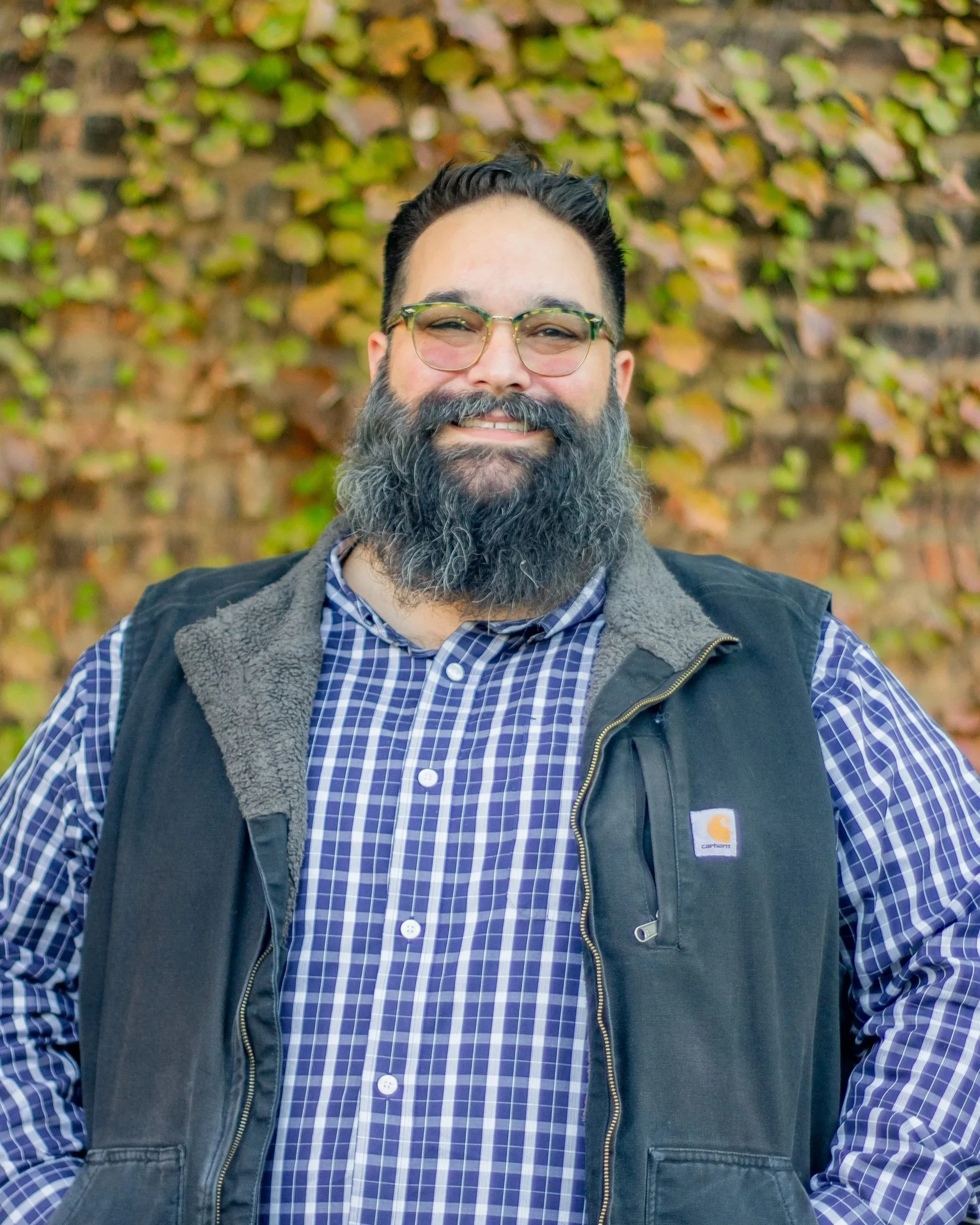 A smiling man with glasses, a dark beard and mustache, wearing a checkered shirt and a black vest, standing outdoors in front of a brick wall covered with green and yellow ivy.