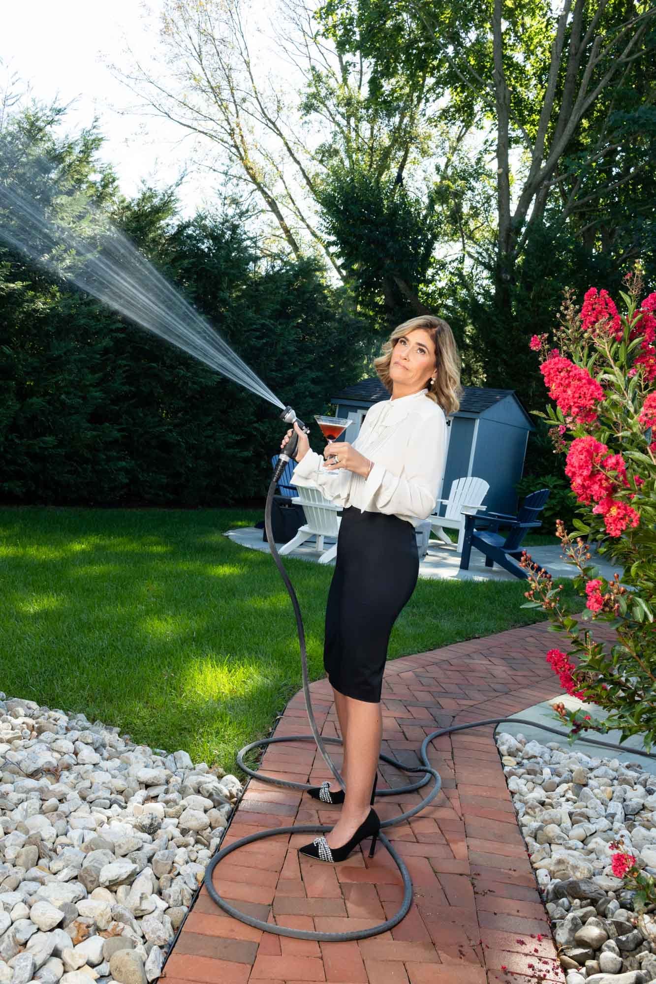A woman in business attire, holding a wine glass and a garden hose with a spray nozzle, stands on a brick pathway in a backyard garden with flowering bushes and trees.