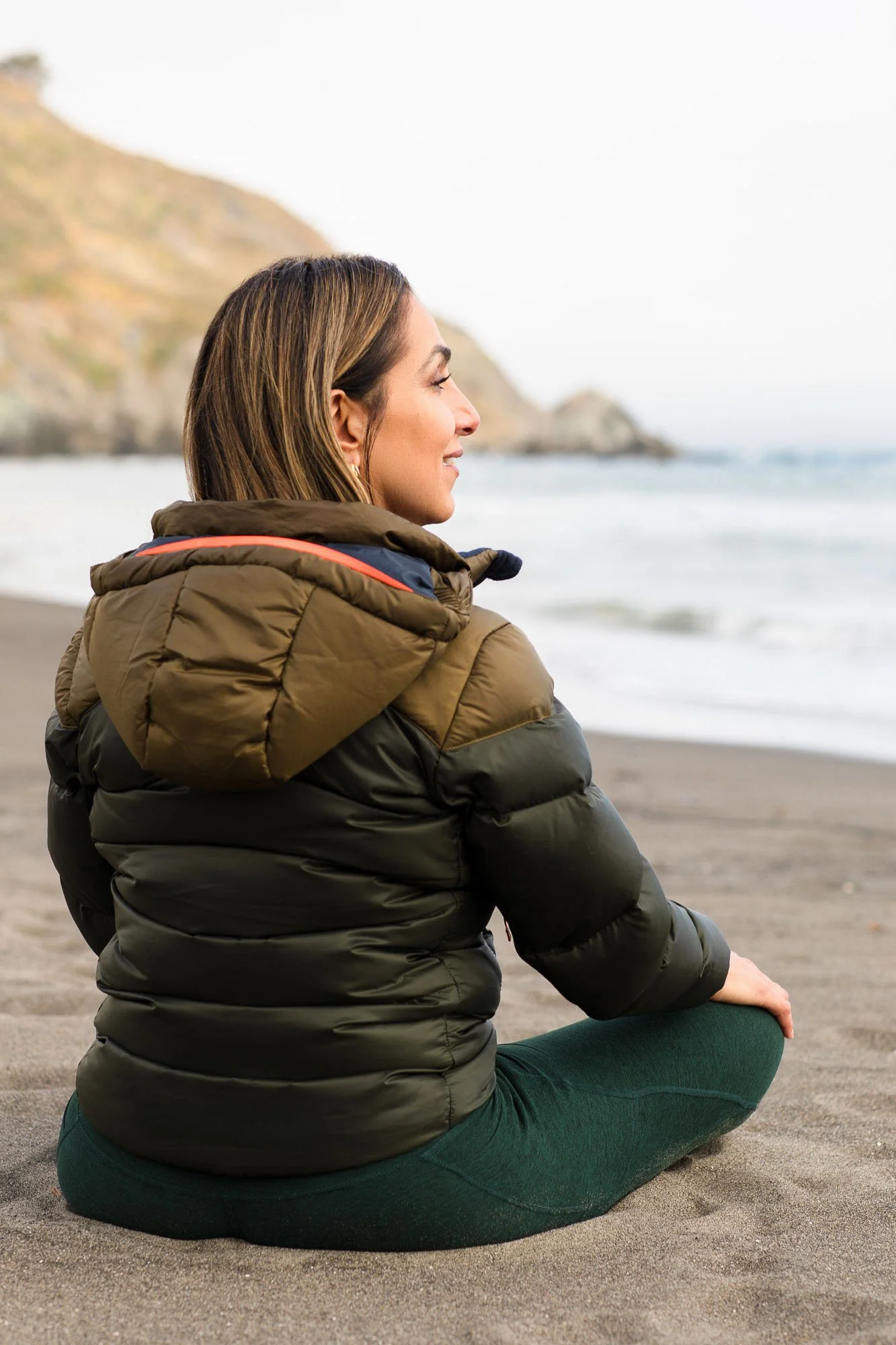 Commercial lifestyle portrait of woman in puffer coat looking at ocean surf on beach, San Francisco Bay Area lifestyle photographer for outdoor travel and apparel brands