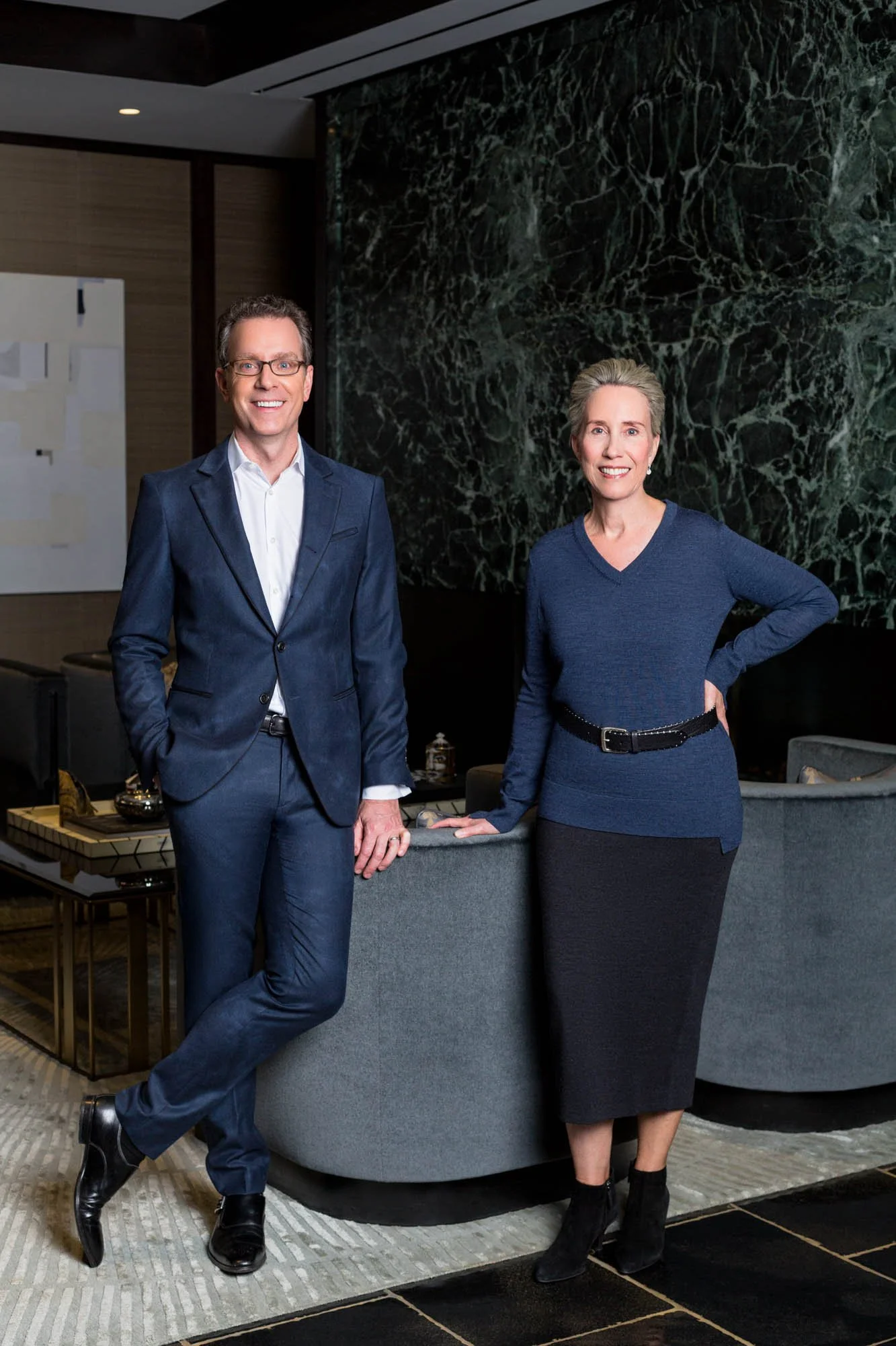 A man in a dark blue suit and a woman in a blue sweater and black skirt standing in a modern office lobby with dark marble wall and contemporary furniture, smiling at the camera.