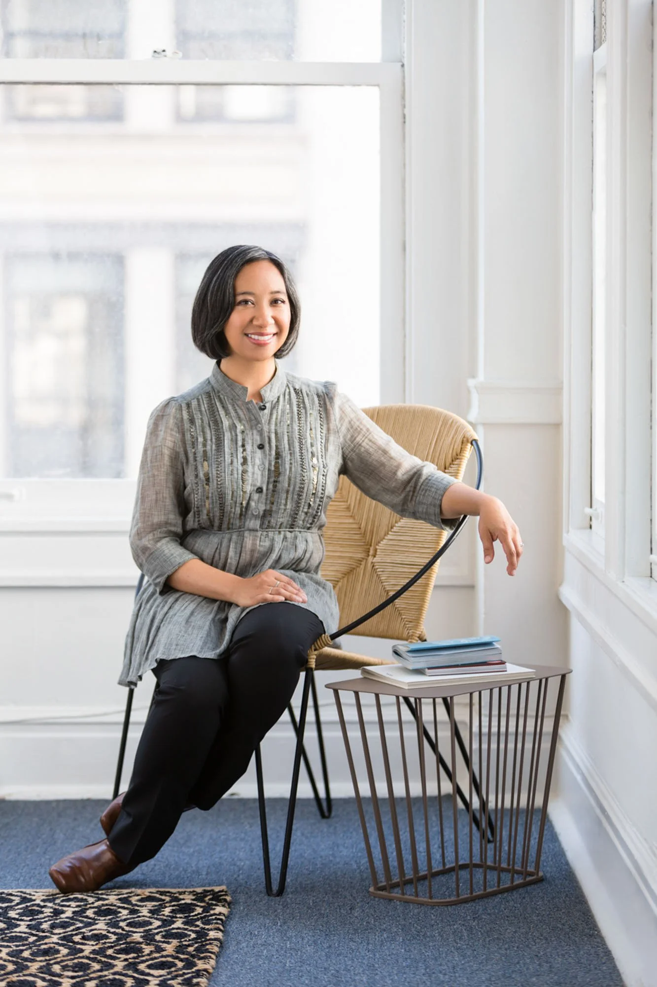 A woman with short dark hair sitting on a modern beige chair by a large window, smiling at the camera, with a small round table holding a few stacked books beside her.