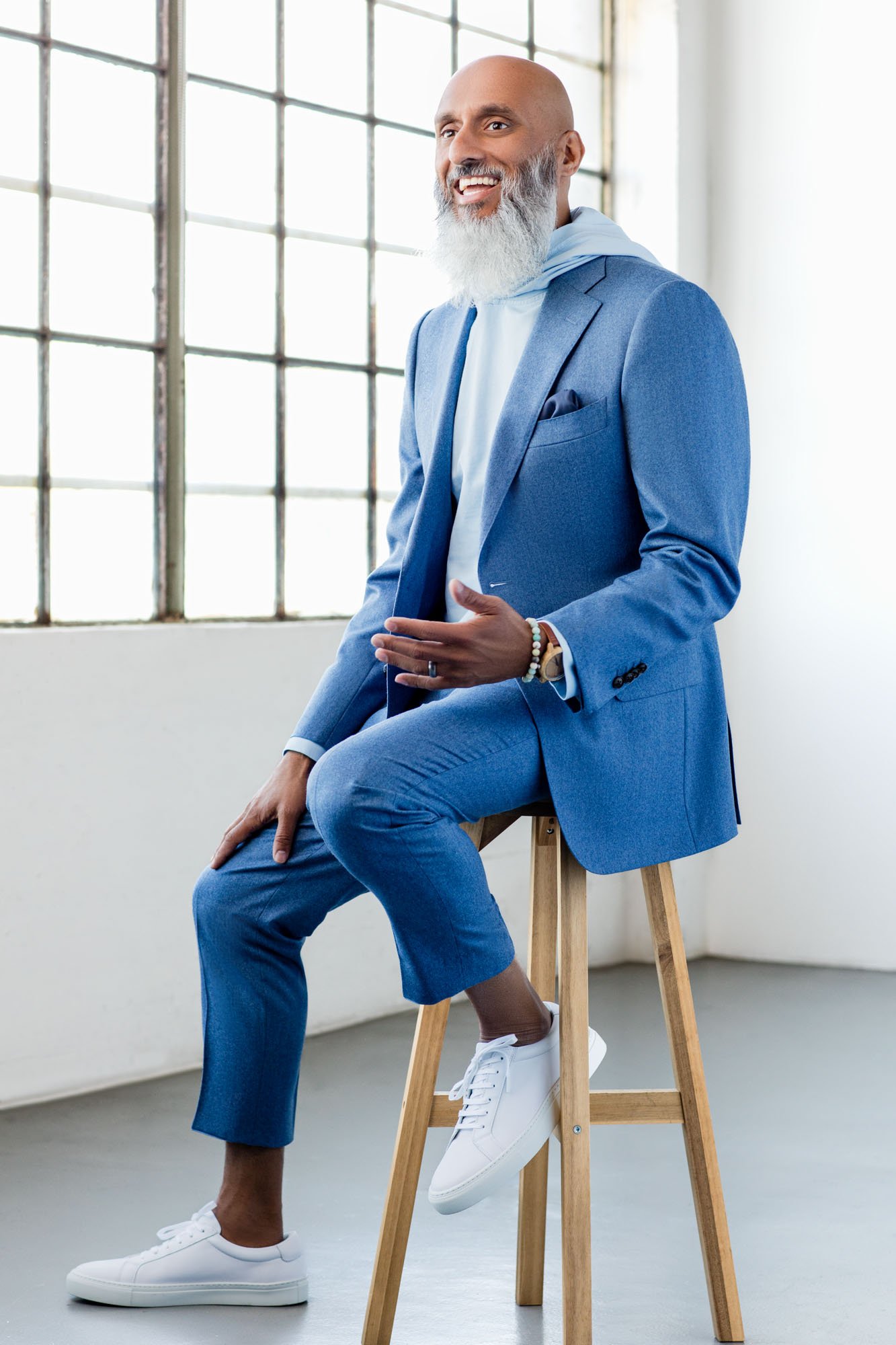 A bald man with a gray beard sitting on a wooden stool in a bright industrial-style room with large windows. He is dressed in a light blue suit with a hoodie underneath, white sneakers, and accessorized with bracelets and a watch, smiling and engagin