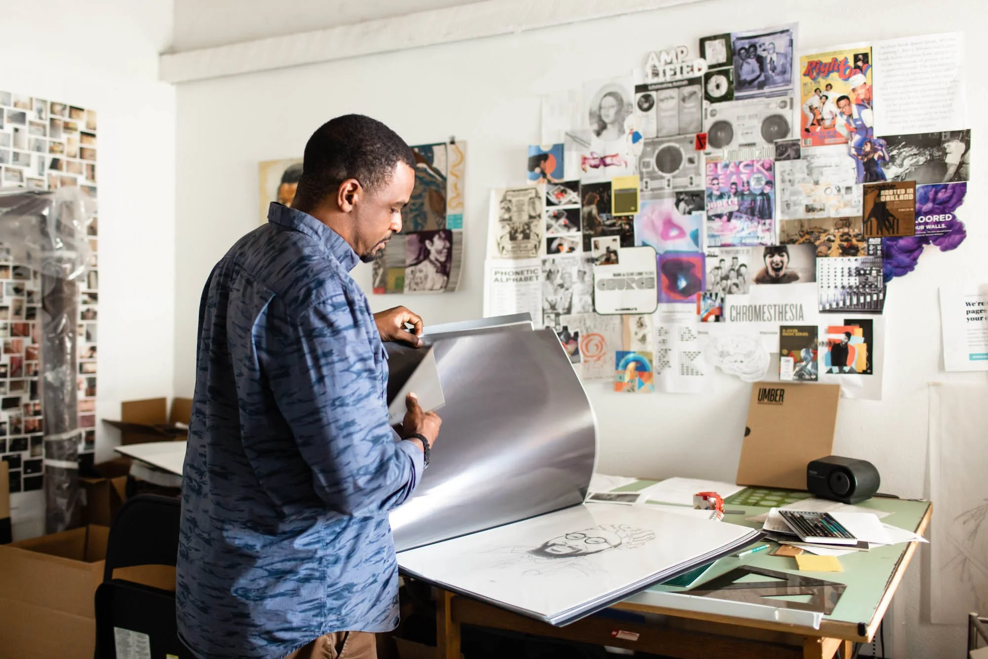 A man in a blue patterned shirt looks through a large binder in an art studio with a collage of artwork on the wall.