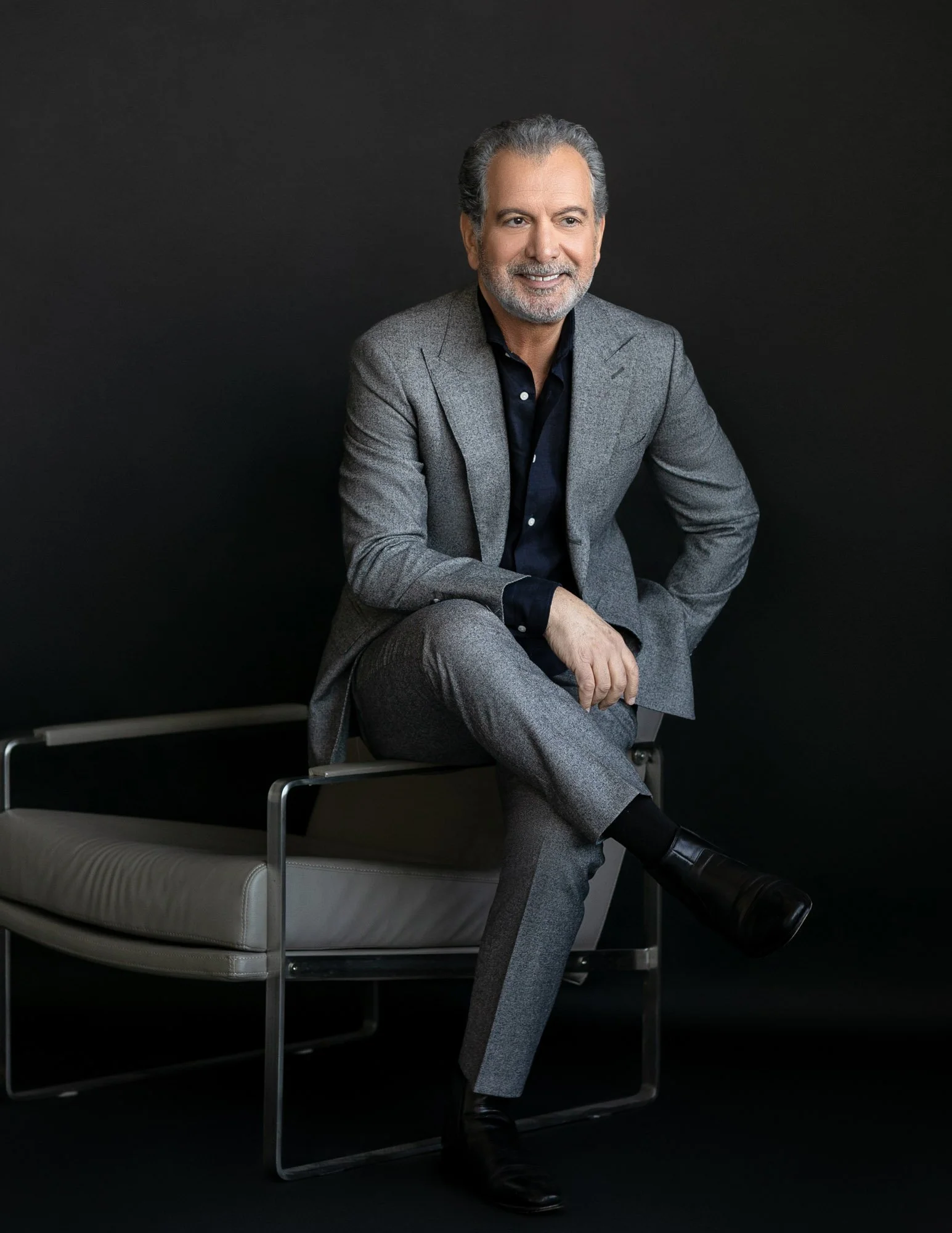 A smiling older man with gray hair and a beard, wearing a gray suit and black shirt, sitting on a modern chair against a black background.