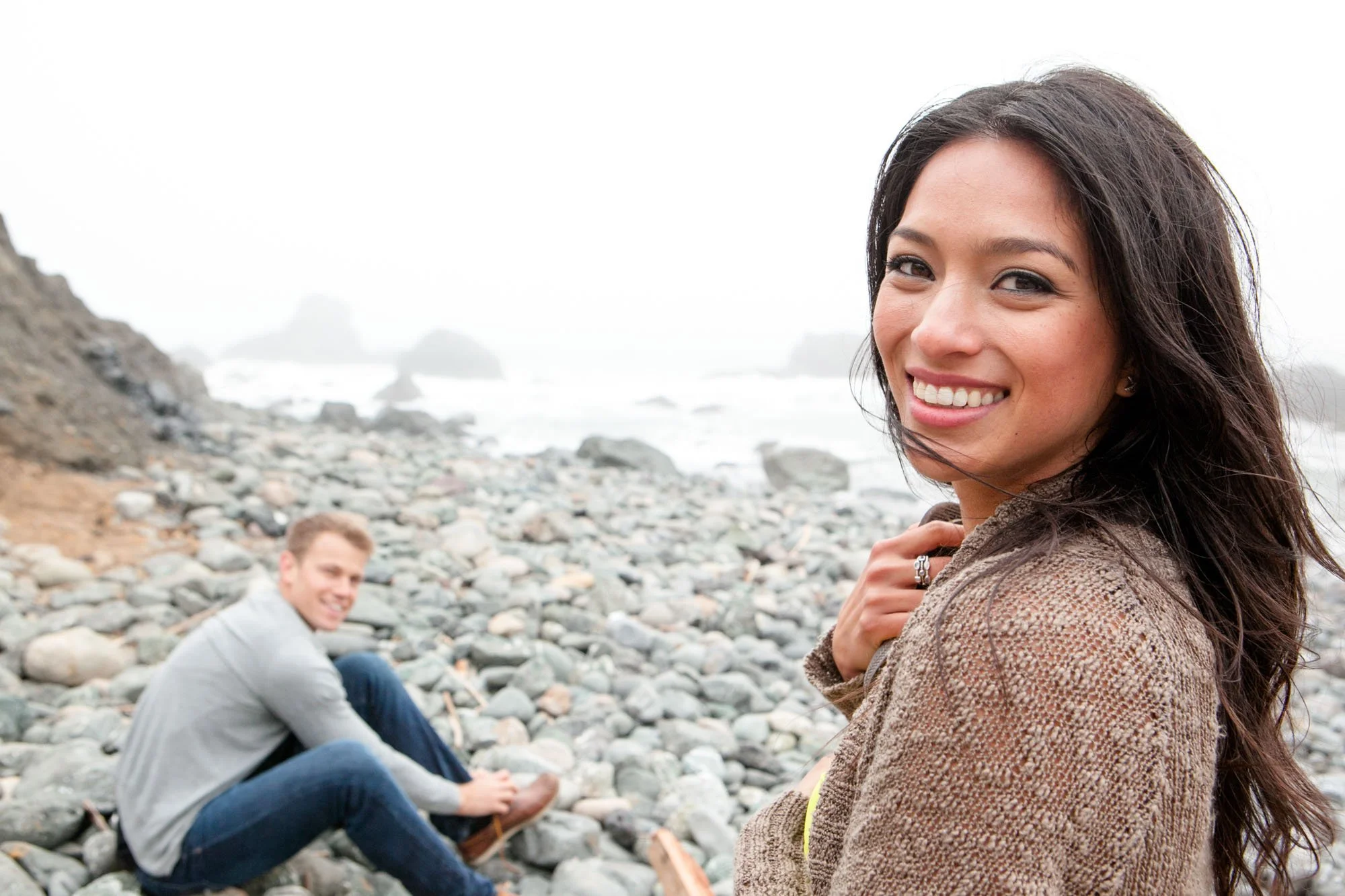 Young woman with long dark hair smiling at the camera on a rocky beach, with a young man sitting on rocks in the background, near the ocean with foggy weather.