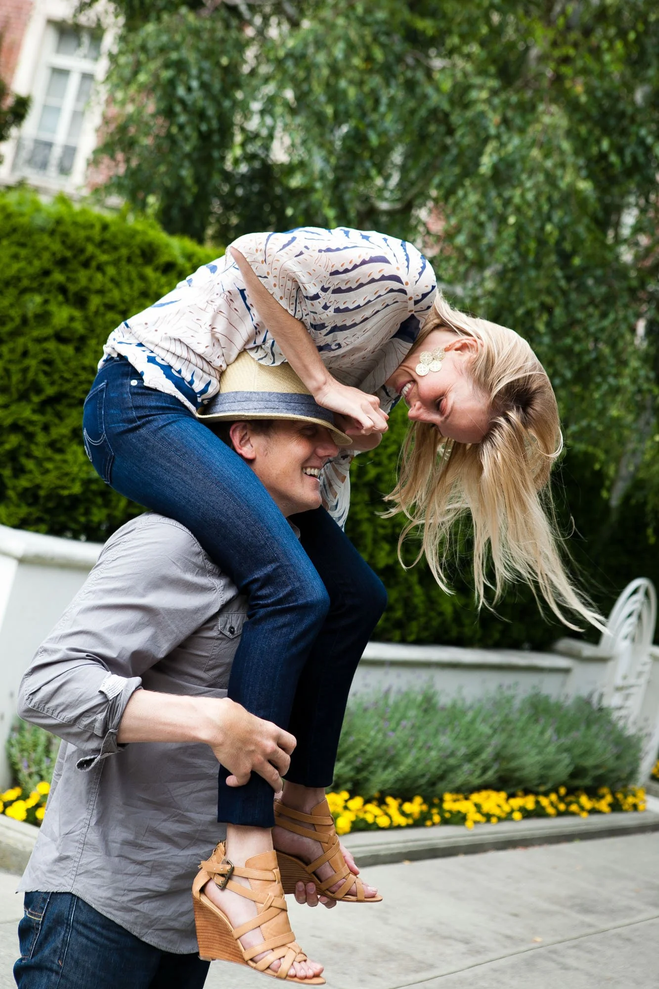 A couple enjoying a playful moment outdoors, with the man giving the woman a piggyback ride. Both are smiling and laughing, surrounded by greenery and flowers.