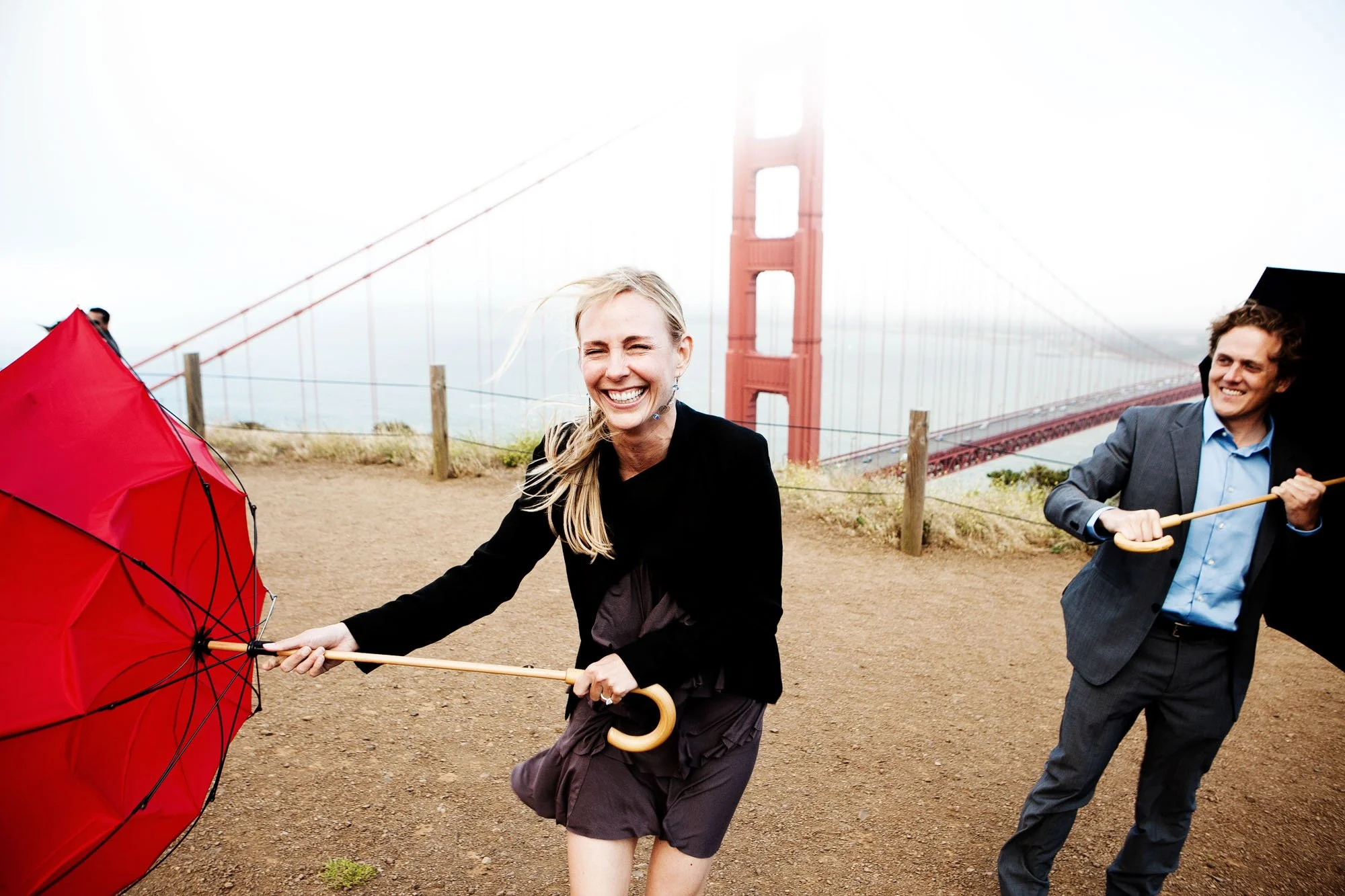 Two people, a smiling woman and a man holding umbrellas, standing on a foggy hill with the Golden Gate Bridge in the background.