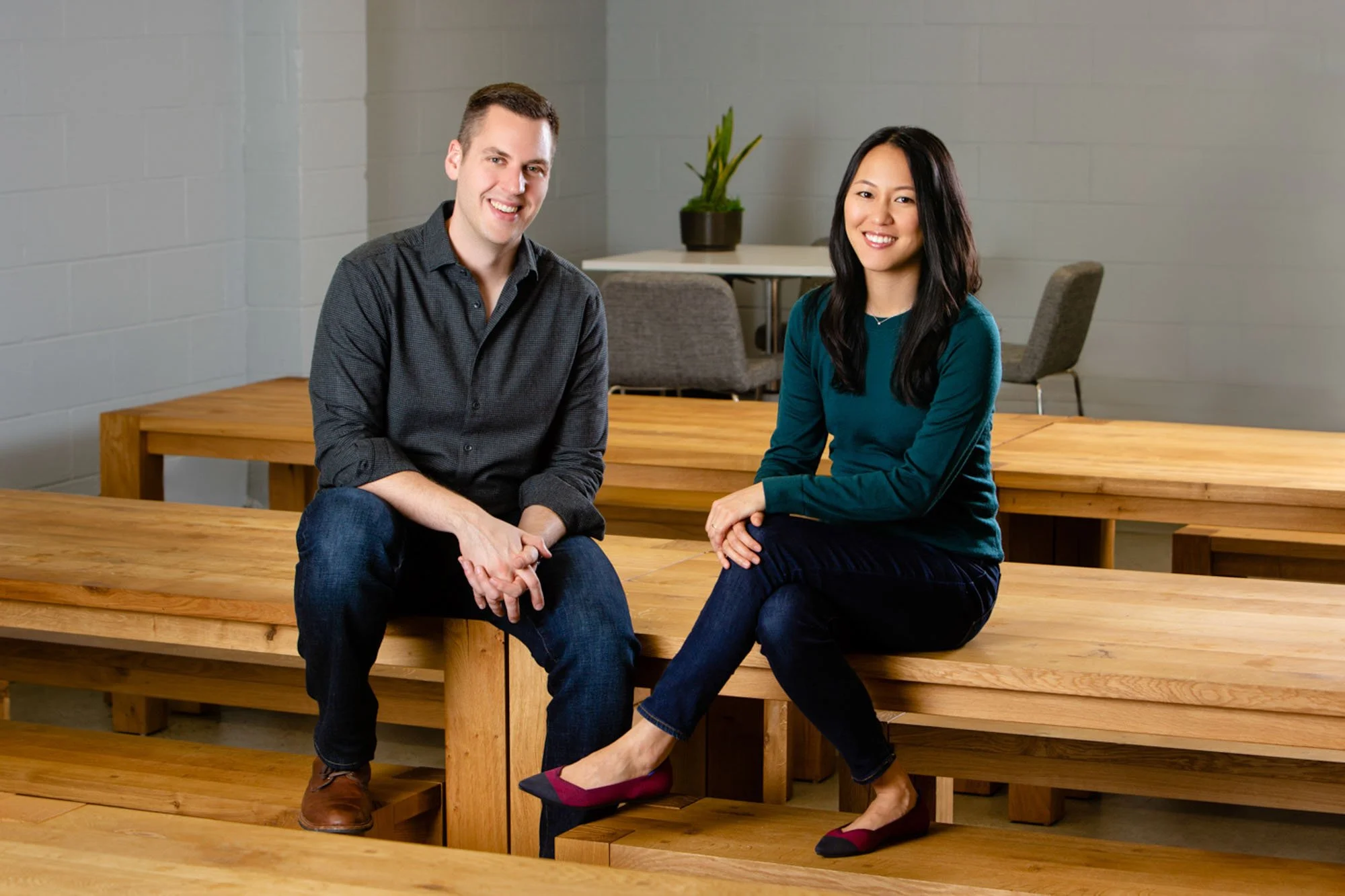 Two people sitting on wooden benches in a room with gray walls, a table with a plant in the background. Commercial portrait and brand photographer in San Francisco and Oakland