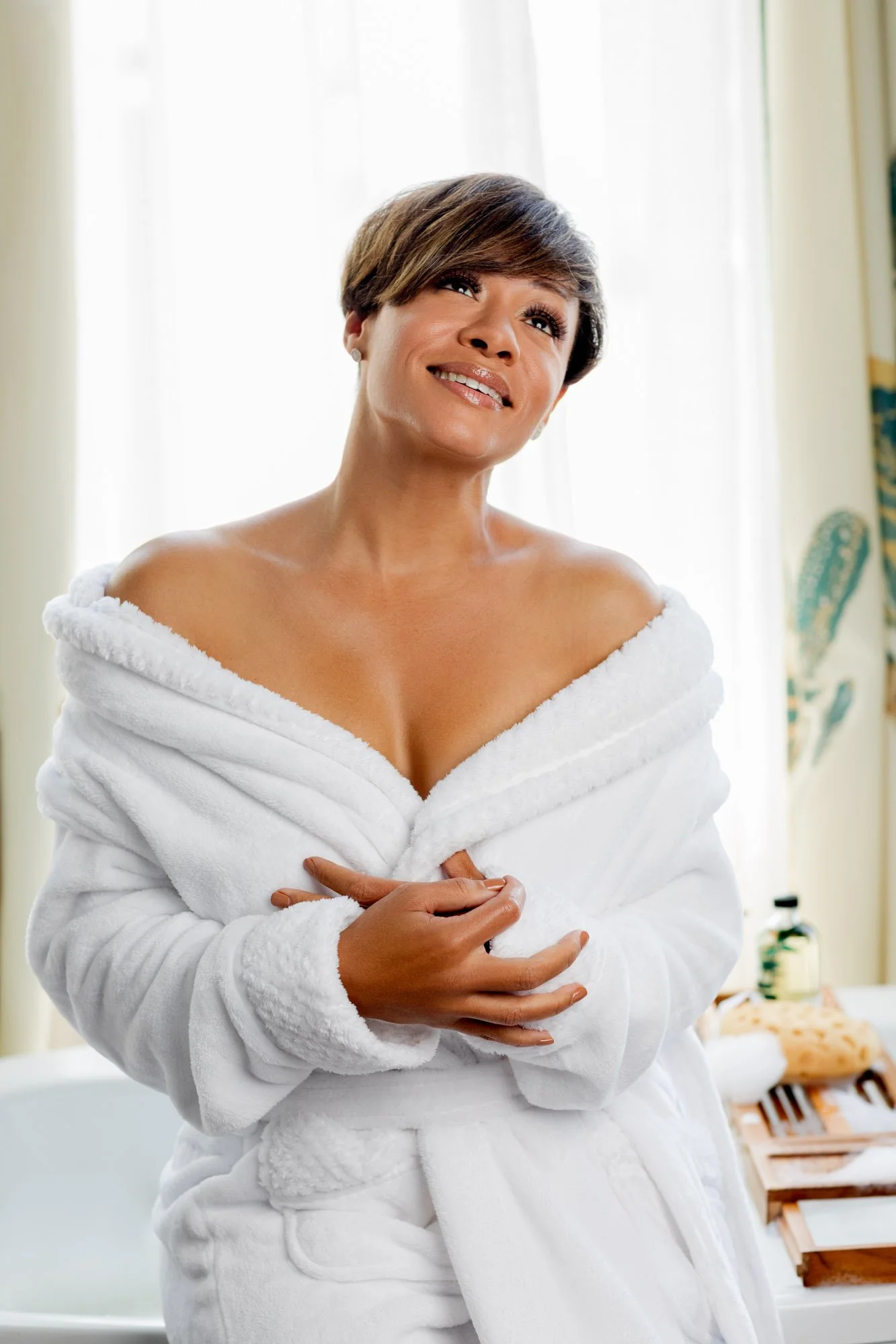 A woman with short hair and earrings wearing a white bathrobe, smiling and holding the bathrobe closed.