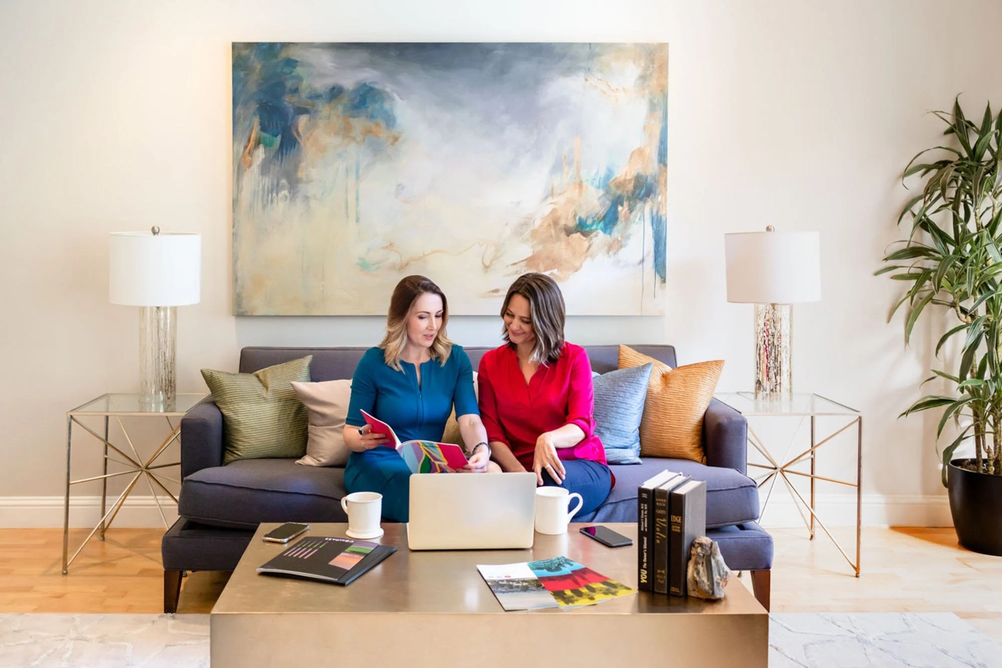 Two women sitting on a sofa in a living room, looking at a laptop and discussing. The sofa has several colorful pillows, and there's a large abstract painting on the wall behind them. A coffee table in front holds books, magazines, a phone, and a mug