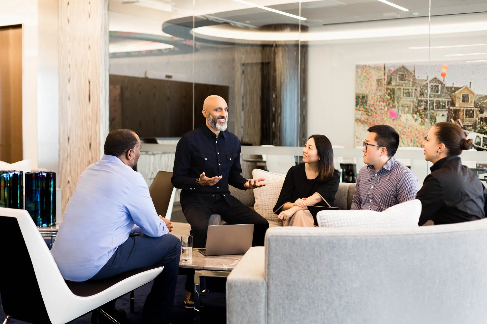 A diverse group of five people having a meeting or discussion in a modern office lounge area, with one man standing and speaking while the others sit on a couch and chairs, engaged and listening.