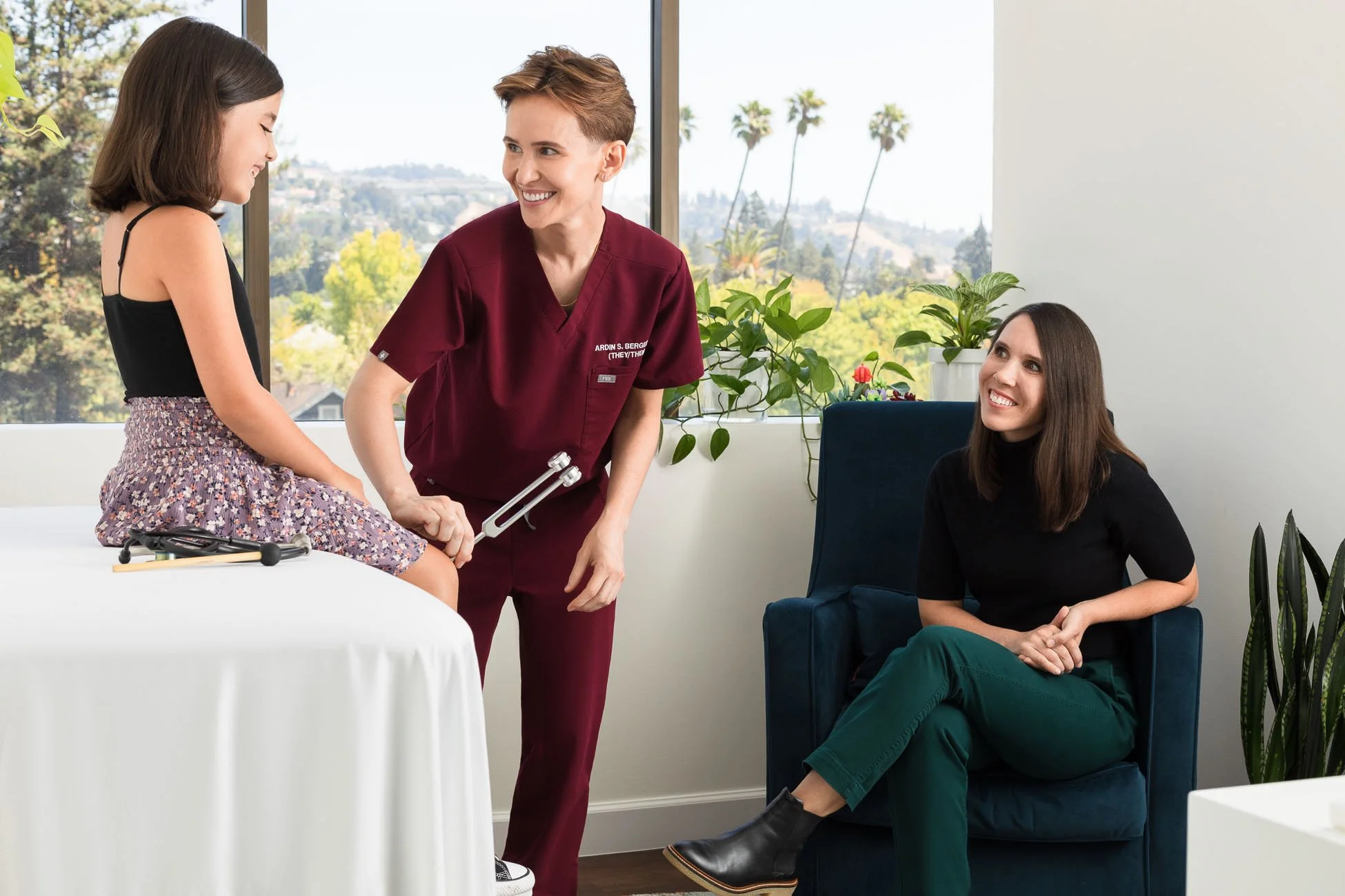 Commercial lifestyle photo of non-binary doctor examining young girl with mother in bright airy medical office, San Francisco Bay Area commercial lifestyle photographer