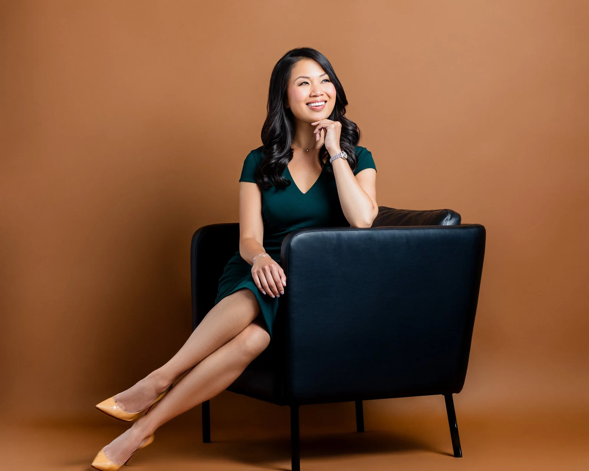 A woman sitting on a black armchair with a brown background, wearing a dark green dress and beige high heels, smiling and resting her chin on her hand.