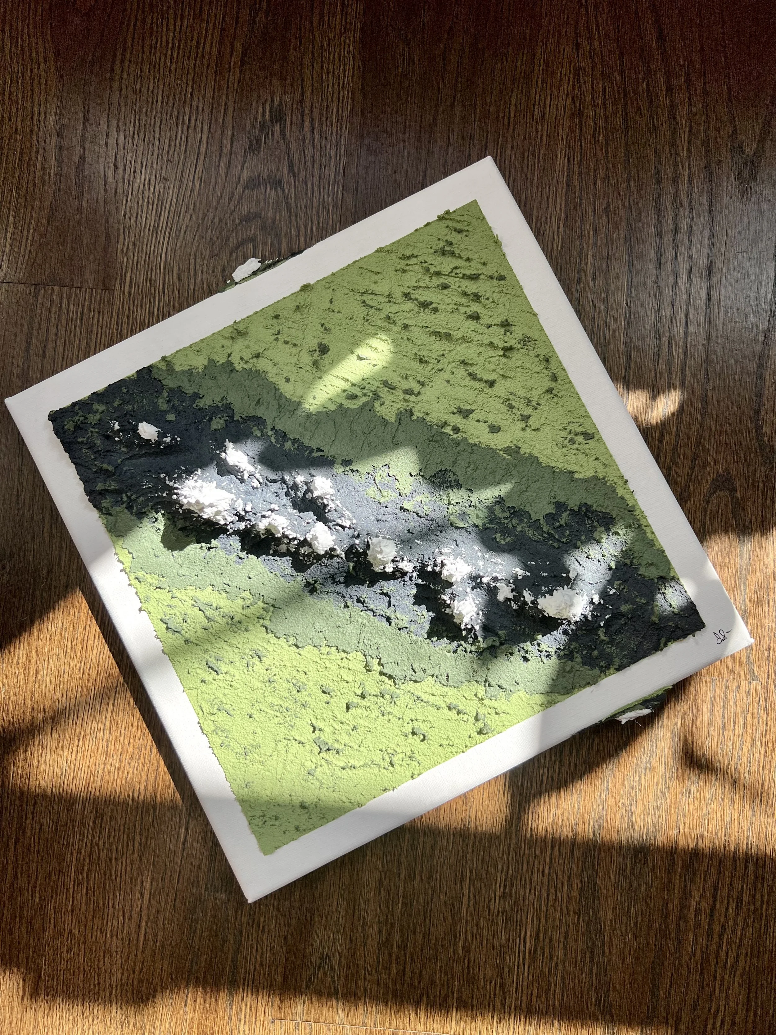 A 3D textured art piece with layered black, green, and white colors on canvas, placed on a wooden surface.