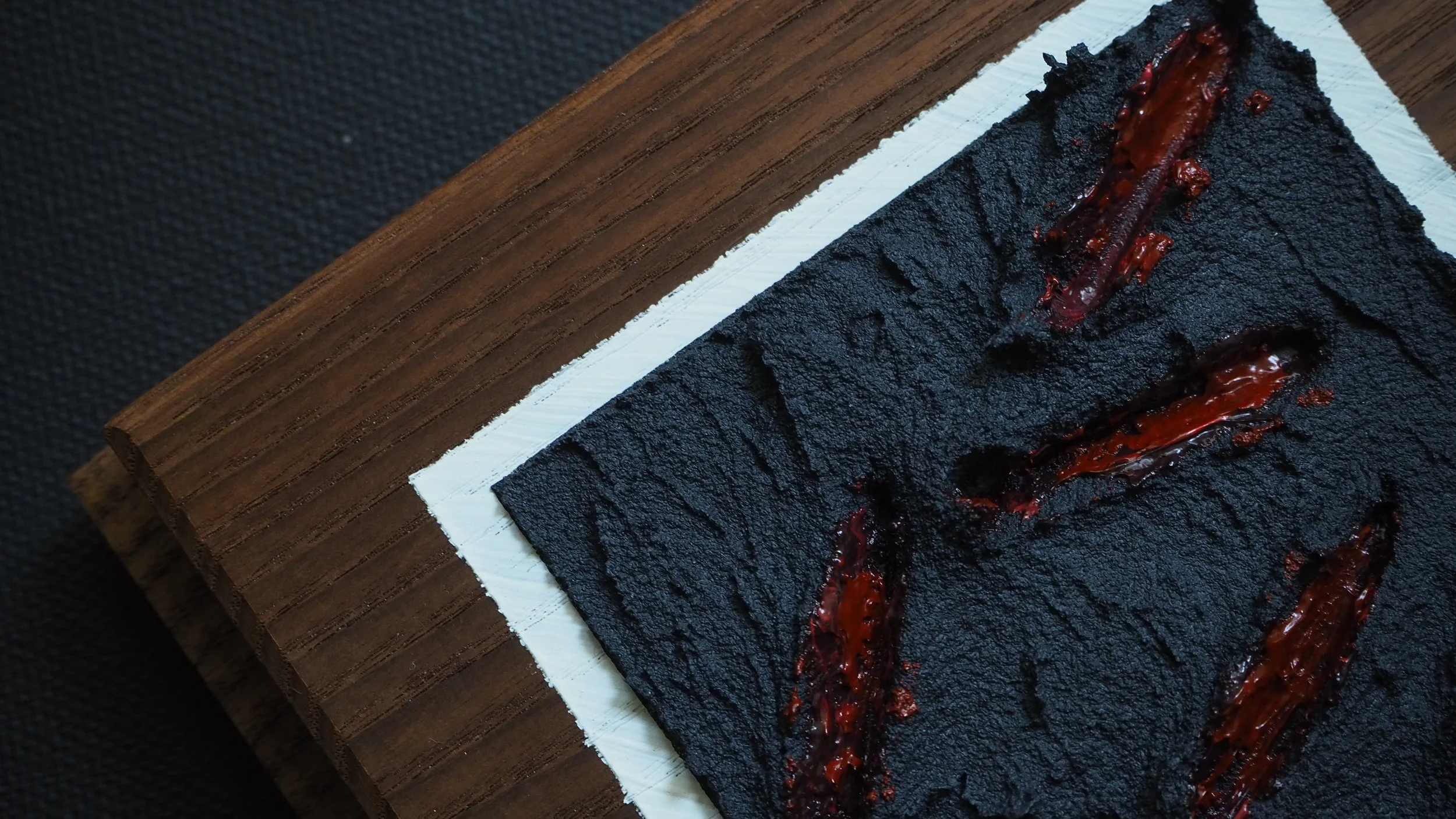 A close-up of a black, crusted rectangular food item with red sauce streaks, placed on parchment paper on a dark wooden surface.