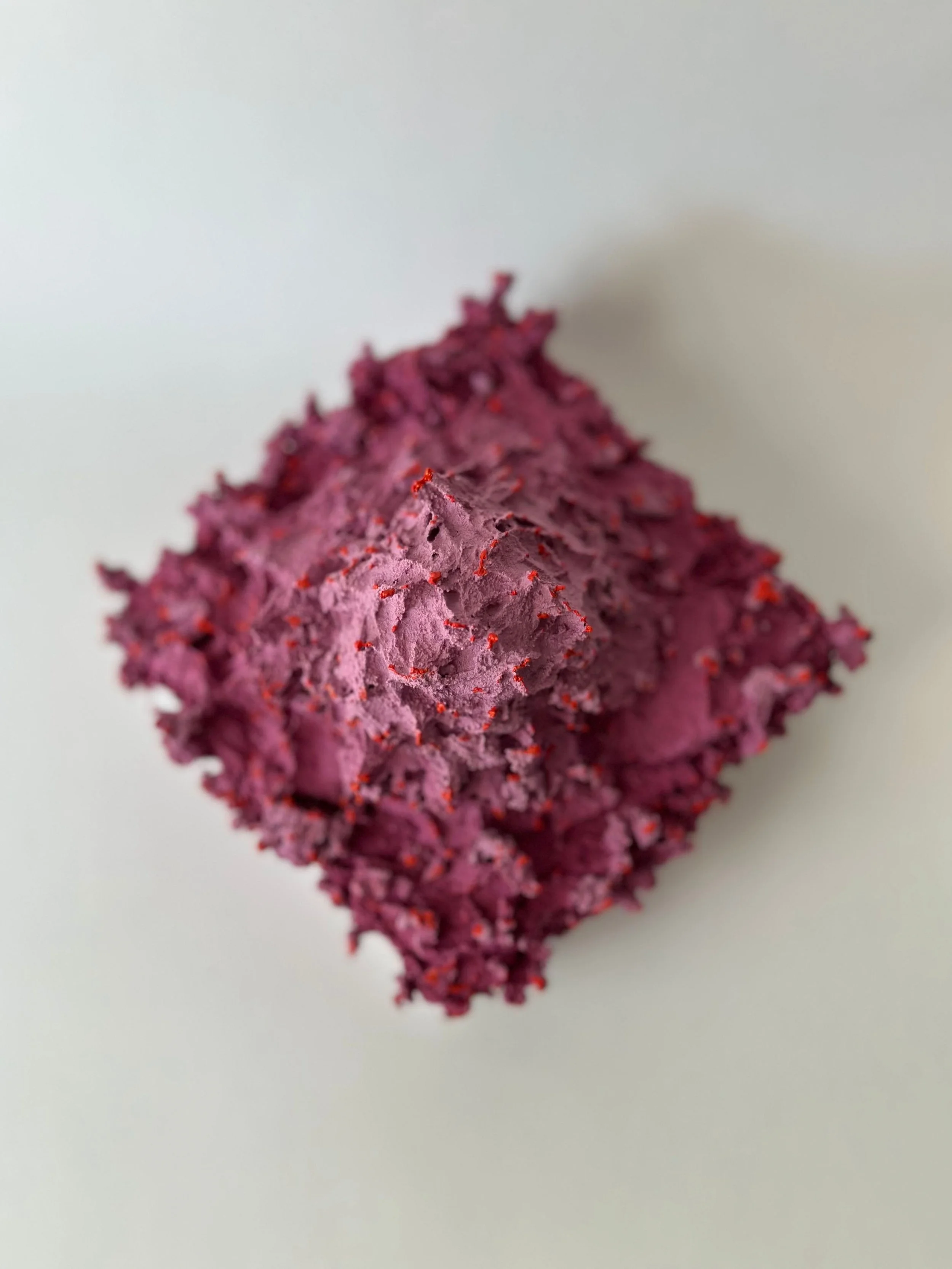 Close-up of a cube-shaped, textured, pinkish-purple object with small red specks on a plain white surface.