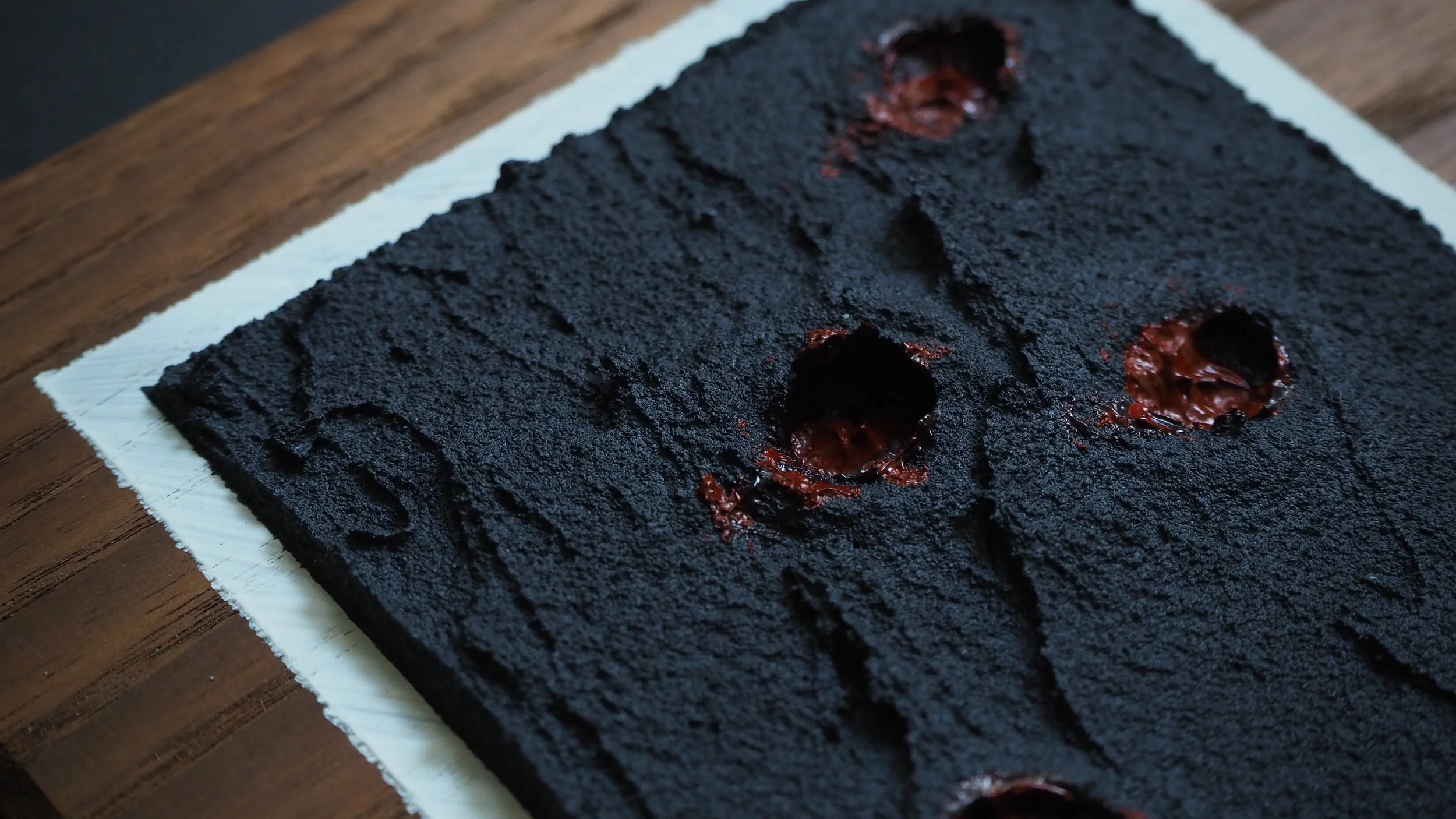 A rectangular, black, textured cake with holes filled with glossy, dark red jelly, resting on white parchment paper on a wooden surface.