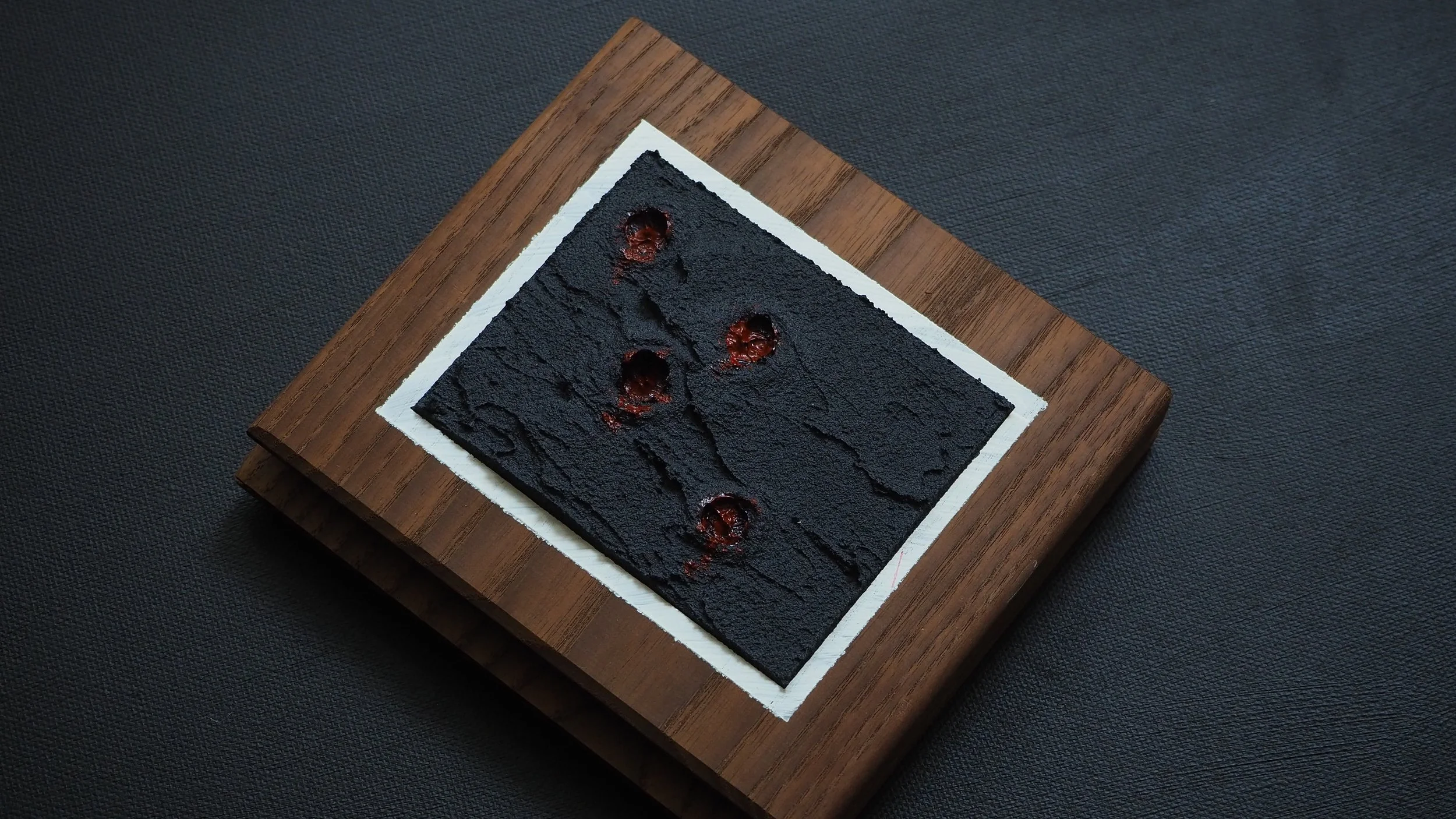 A black textured square dessert with red streaks and holes on a white paper on a wooden board.