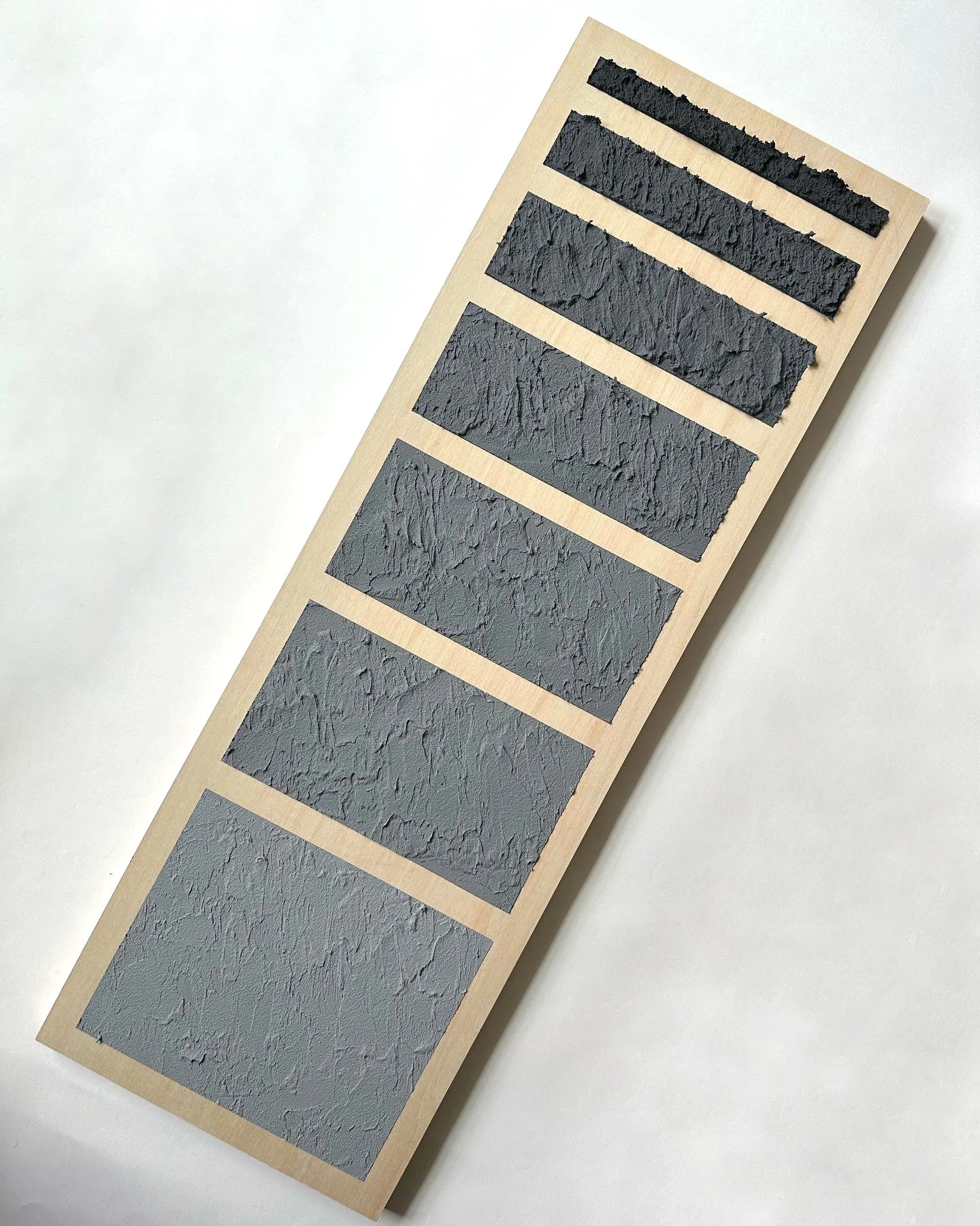 A wooden display board with six different shades of gray paint or plaster samples, each with a textured, rough surface.