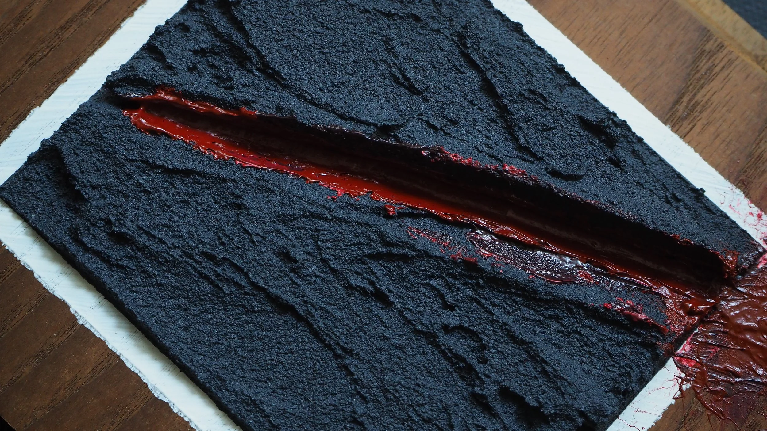 A black layered cake with a dark red filling, textured black surface, on a white board on a wooden table.