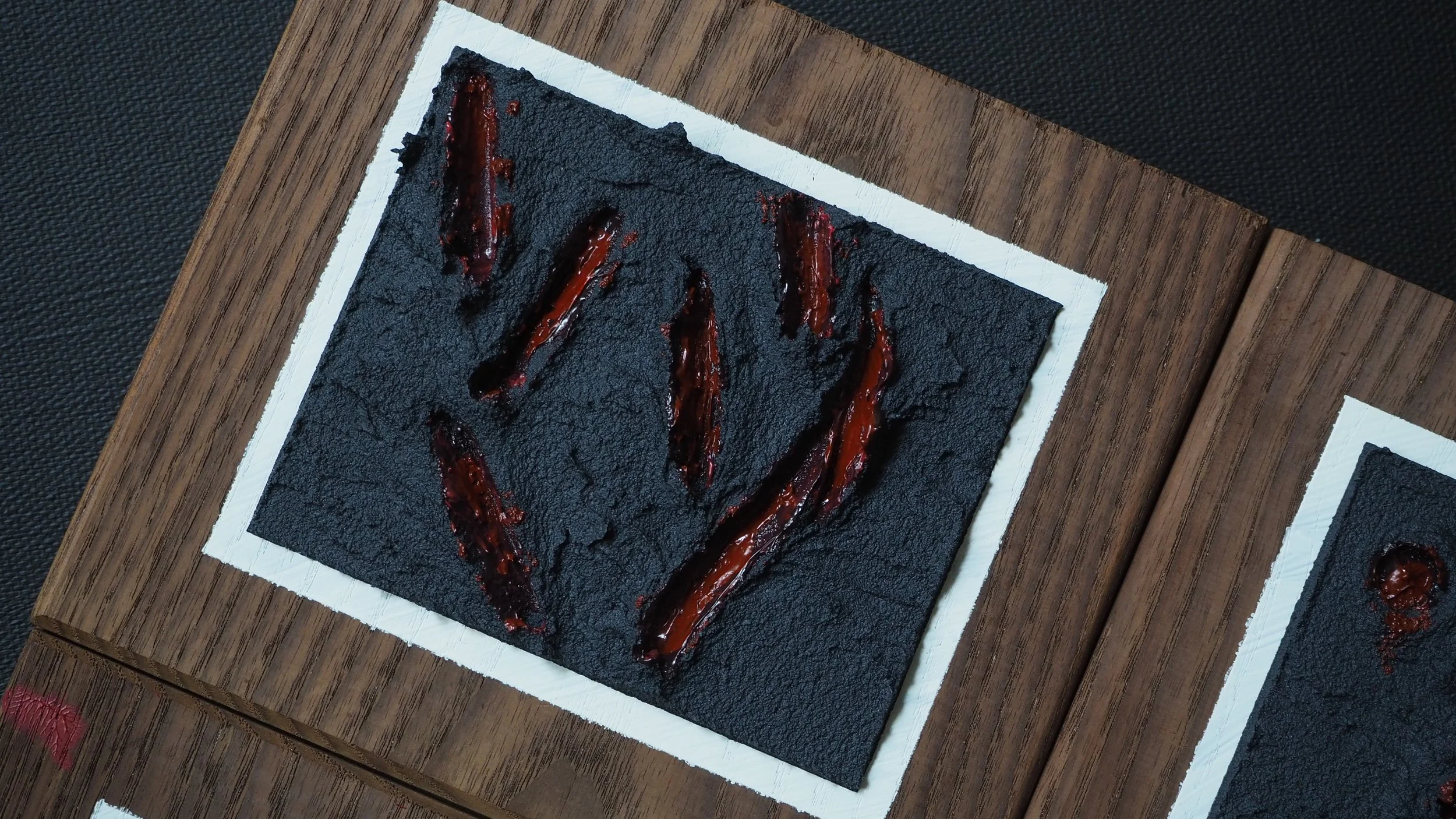 An art piece on a black textured background with red glossy streaks forming a symbol, mounted on a wooden surface with a white border.