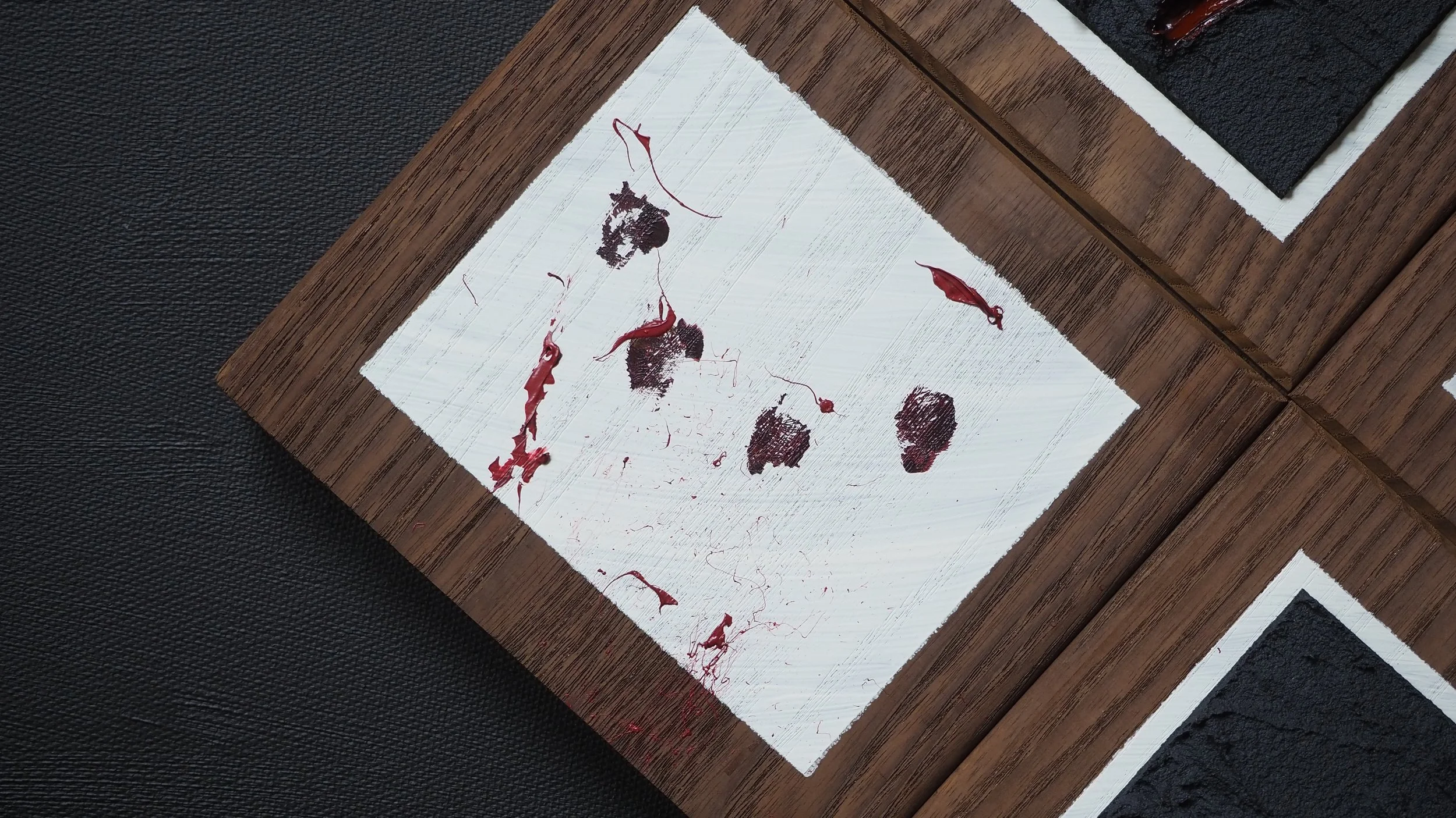 A white piece of paper with black smeared ink and red paint drips, set on a wooden surface with dark textured backgrounds.