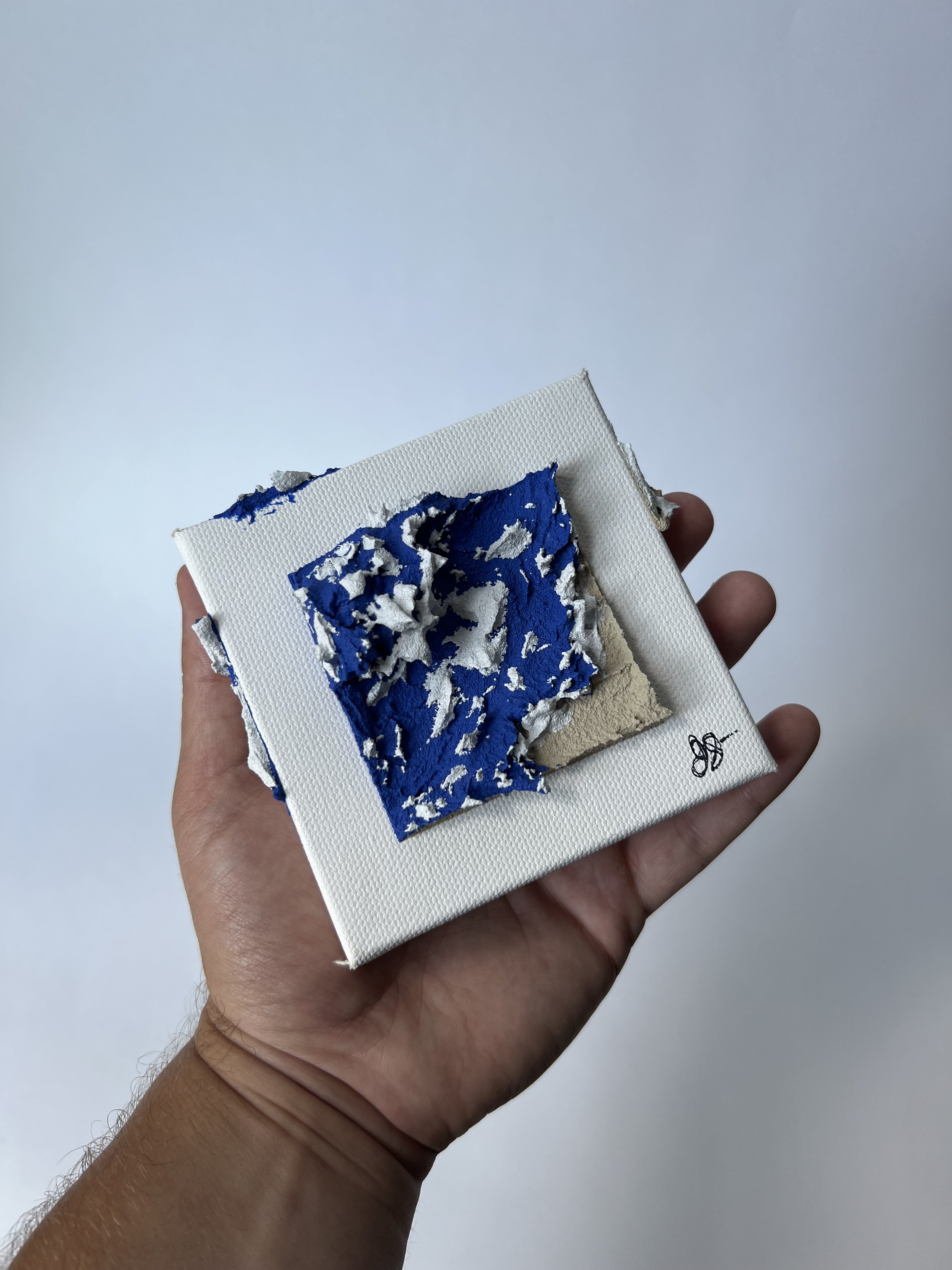 A hand holding a small white canvas with torn, layered paper pieces in blue, white, and beige colors.