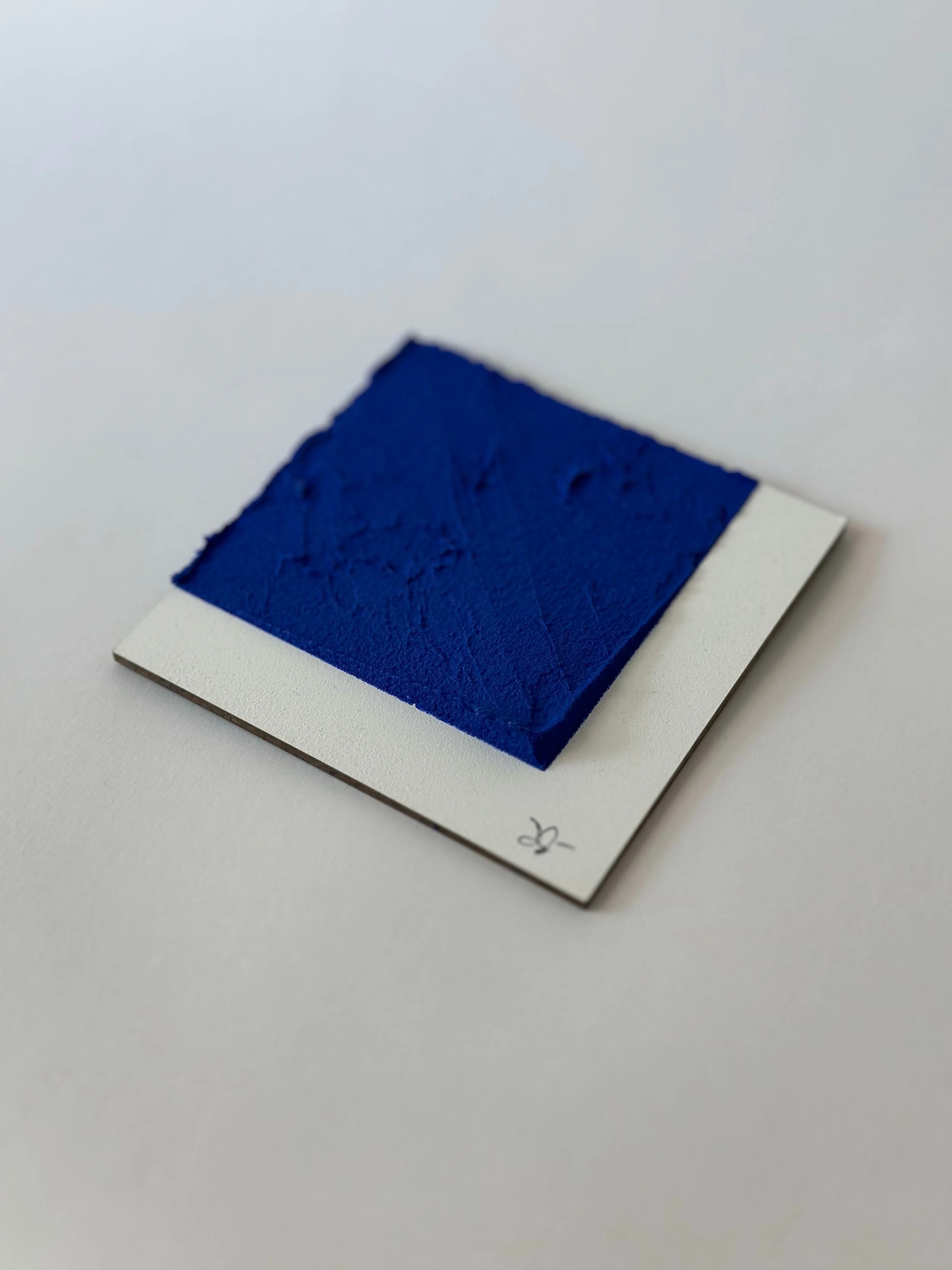 Square swatch of textured blue paint on a white sample card with handwritten markings.