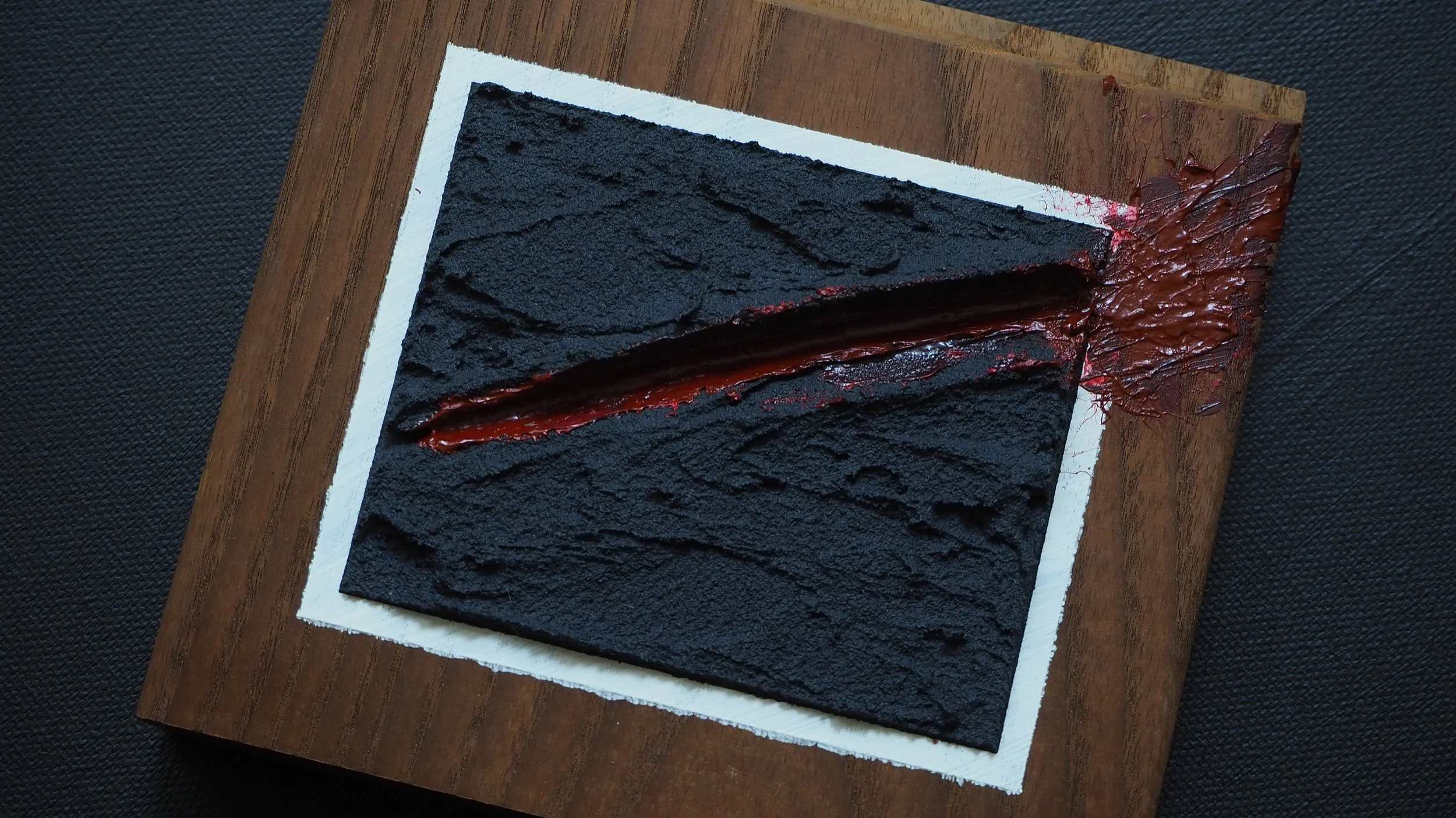 A slice of black cake with a red filling on a white and wooden tray, with some red glaze and textured surface.