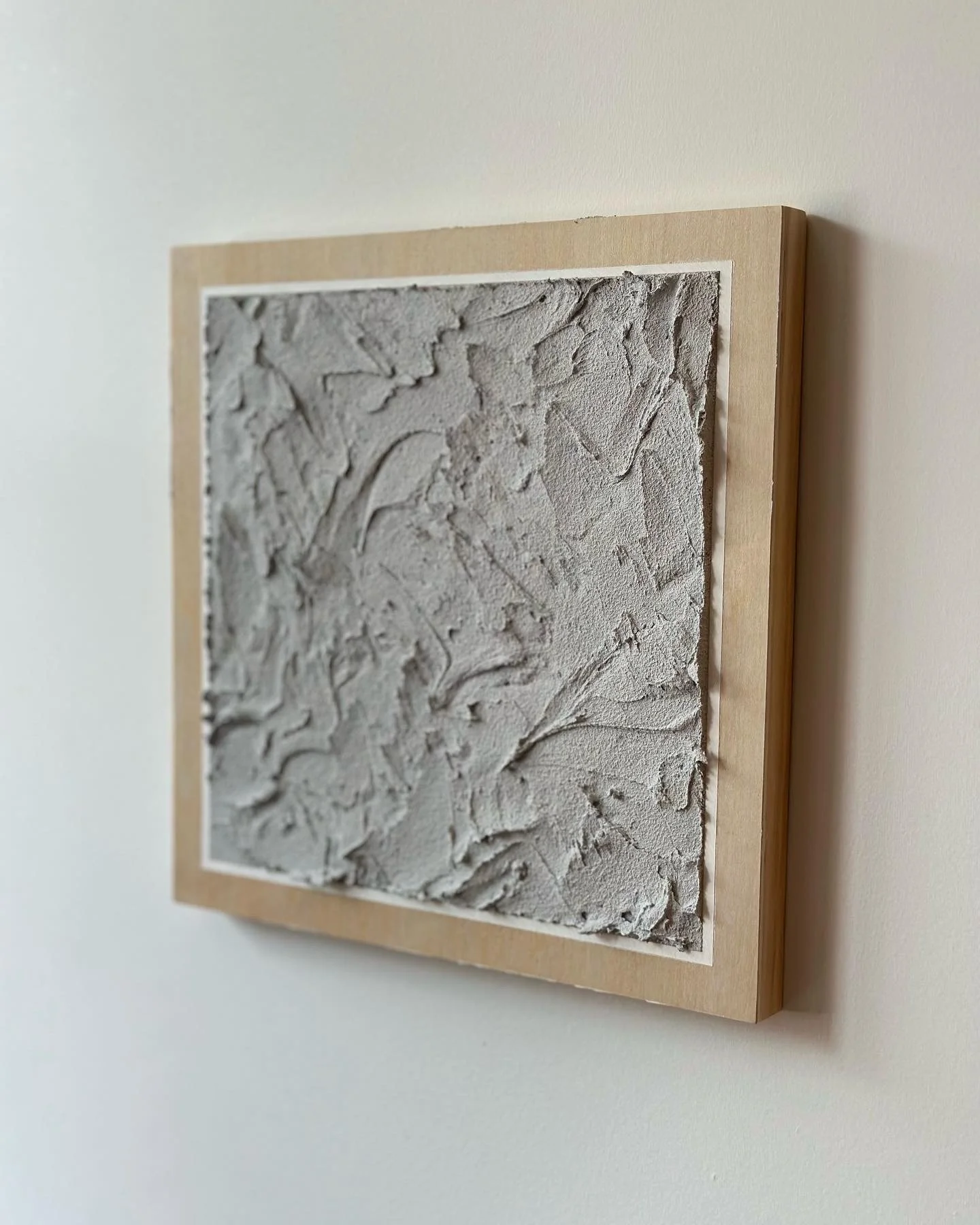 Textured gray artwork framed in light wood hanging on a white wall.
