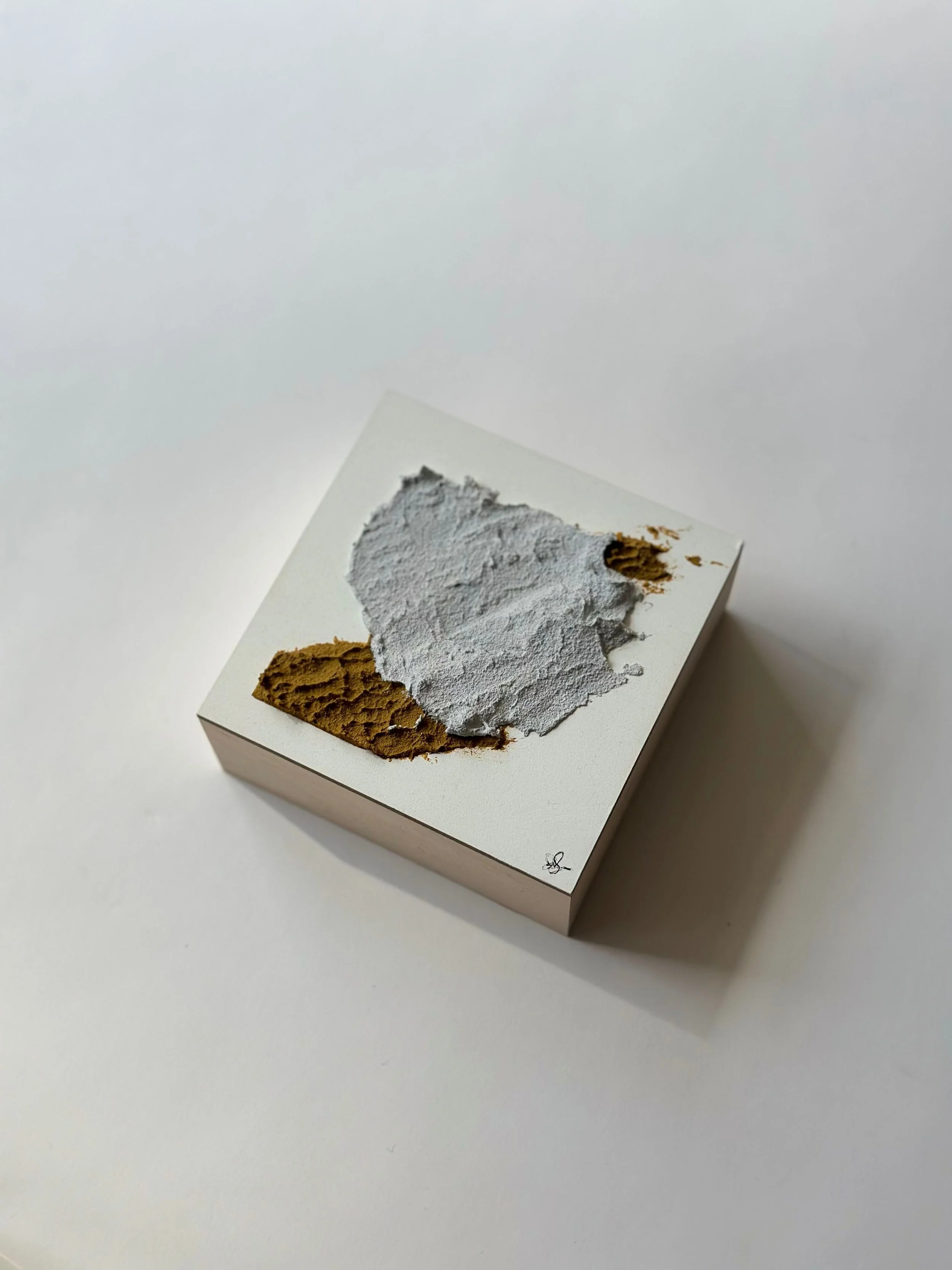 Square white box with textured gray and brown material on top, likely a cosmetic or skincare product, on a plain white background.