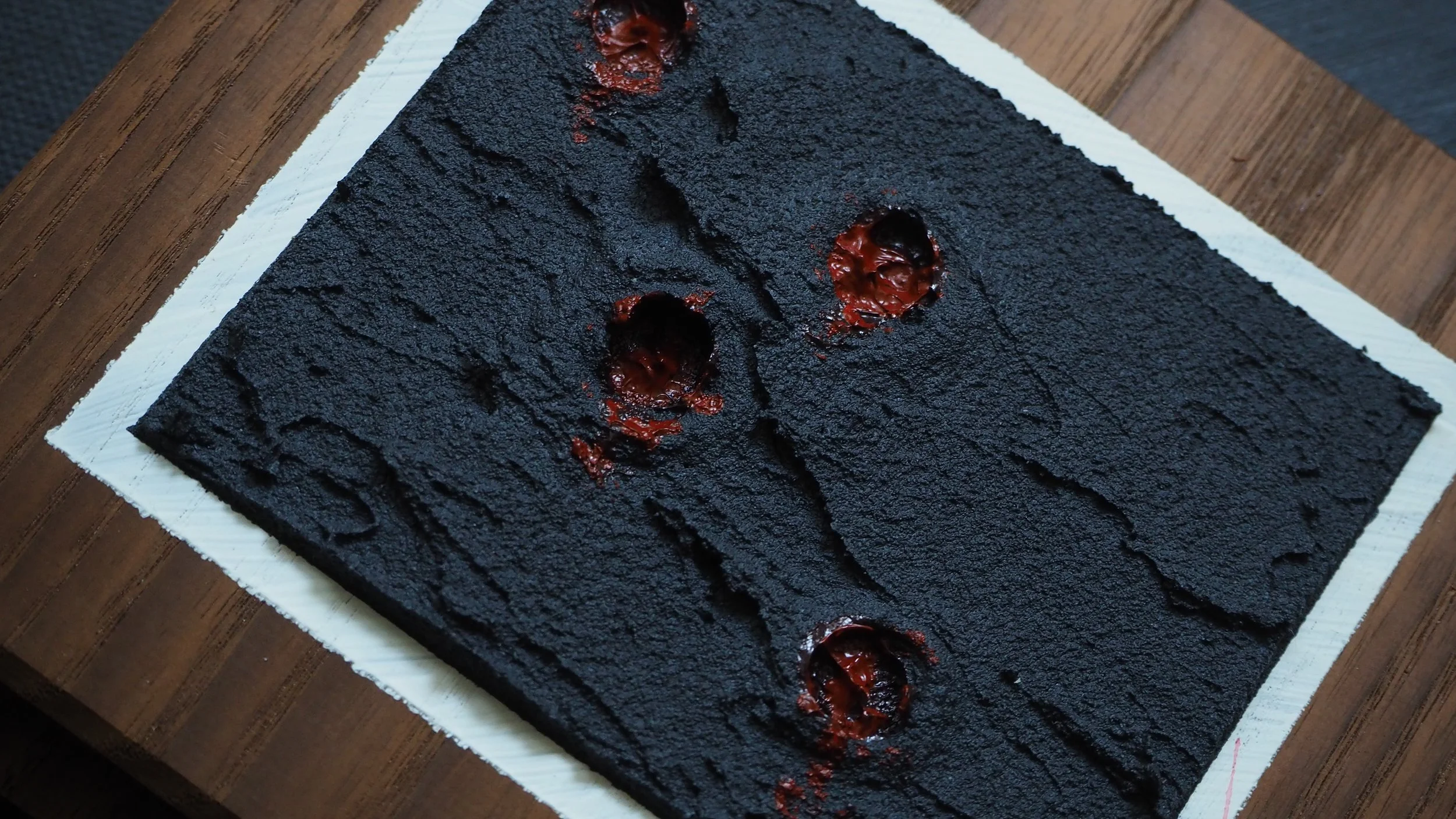 A textured black dessert with three small holes filled with red jelly, placed on white paper on a wooden surface.