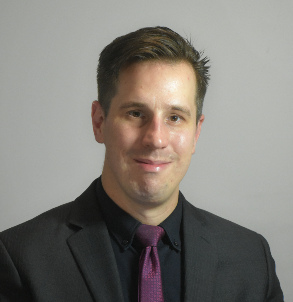 Professional young man in a black suit, dark dress shirt, and purple tie, smiling against a plain background.