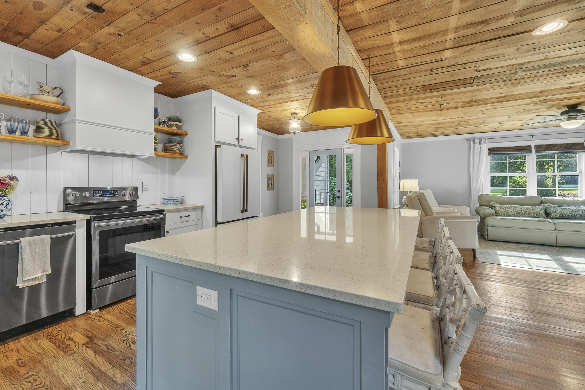 Open-concept kitchen with white cabinetry, stainless steel appliances, light-colored countertops, and open shelving with dishes and glasses. A large island with a light blue base and white countertop is in the foreground. The ceiling is wood-paneled 