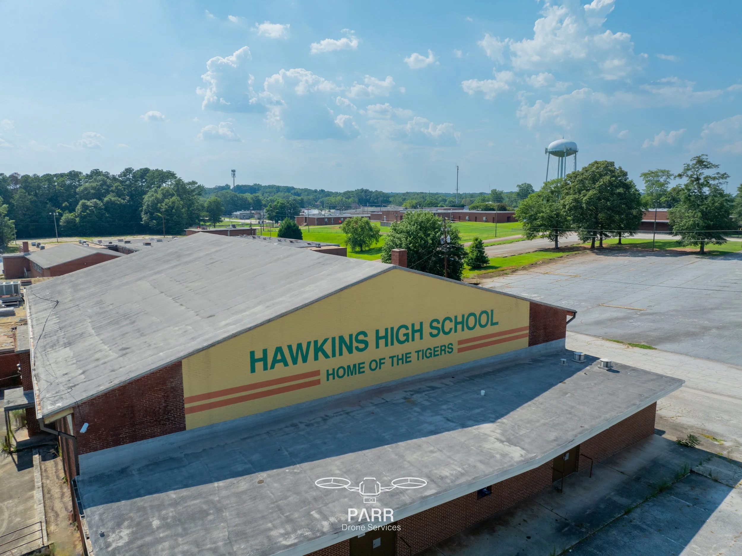 Drone view of Hawkins High School building with a large painted sign on the side that reads 'Hawkins High School, Home of the Tigers.' The school is surrounded by parking lots, trees, and open fields under a partly cloudy sky.