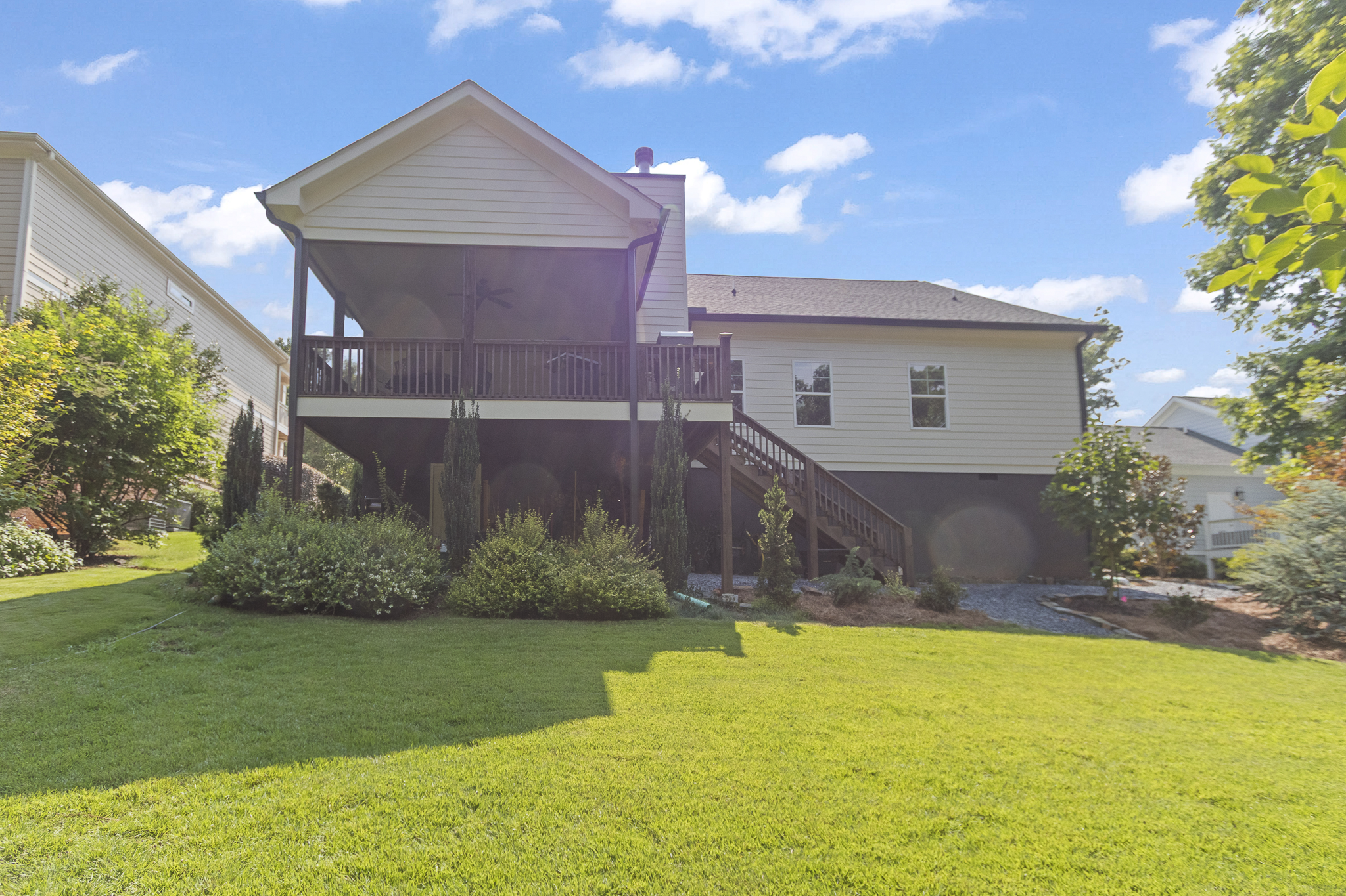 Back view of a two-story house with a wooden deck and stairs, surrounded by a green lawn and trees on a sunny day with a partly cloudy sky.