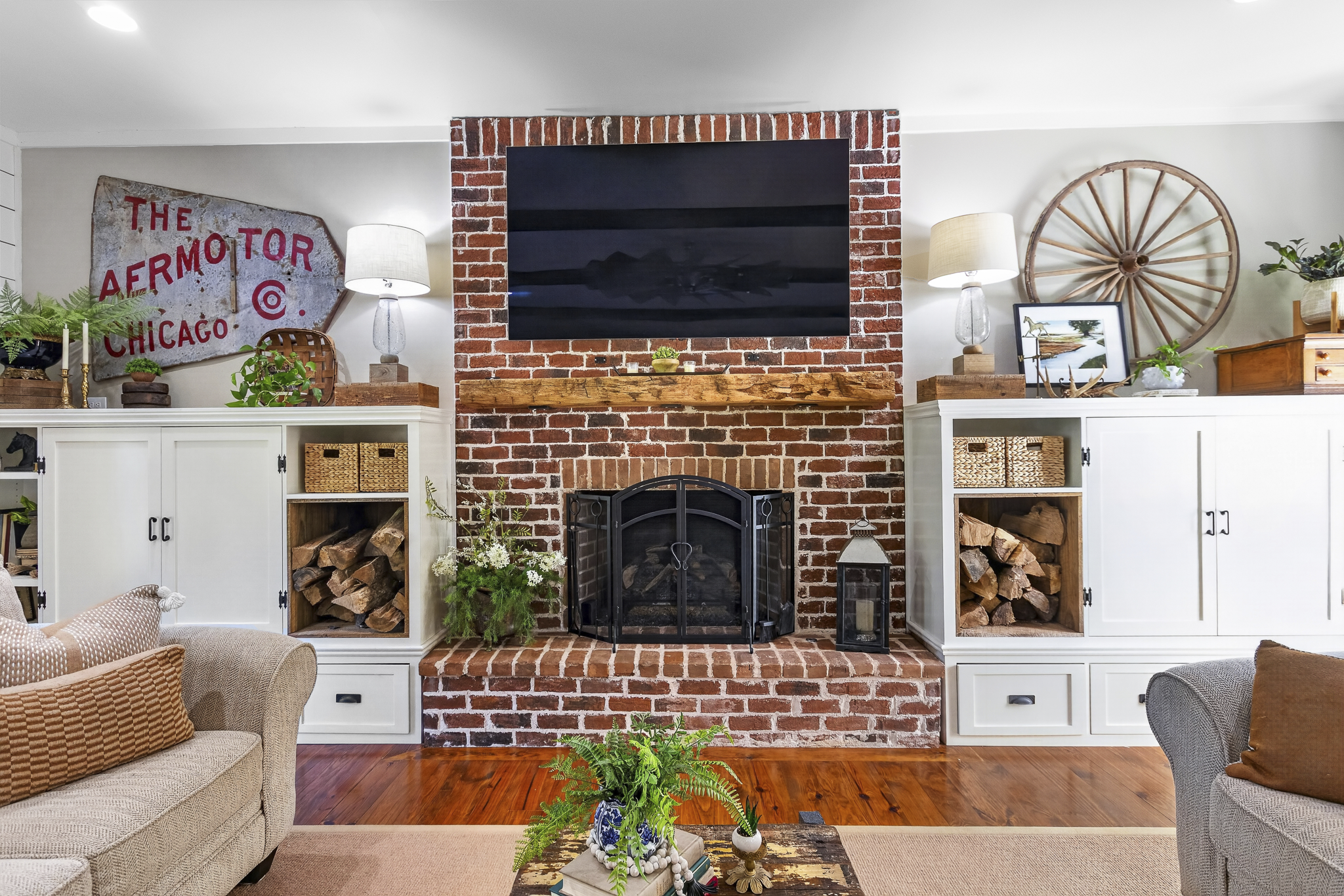 Living room with brick fireplace, mounted TV, white cabinets, and decorative items including plants, framed pictures, and lanterns.