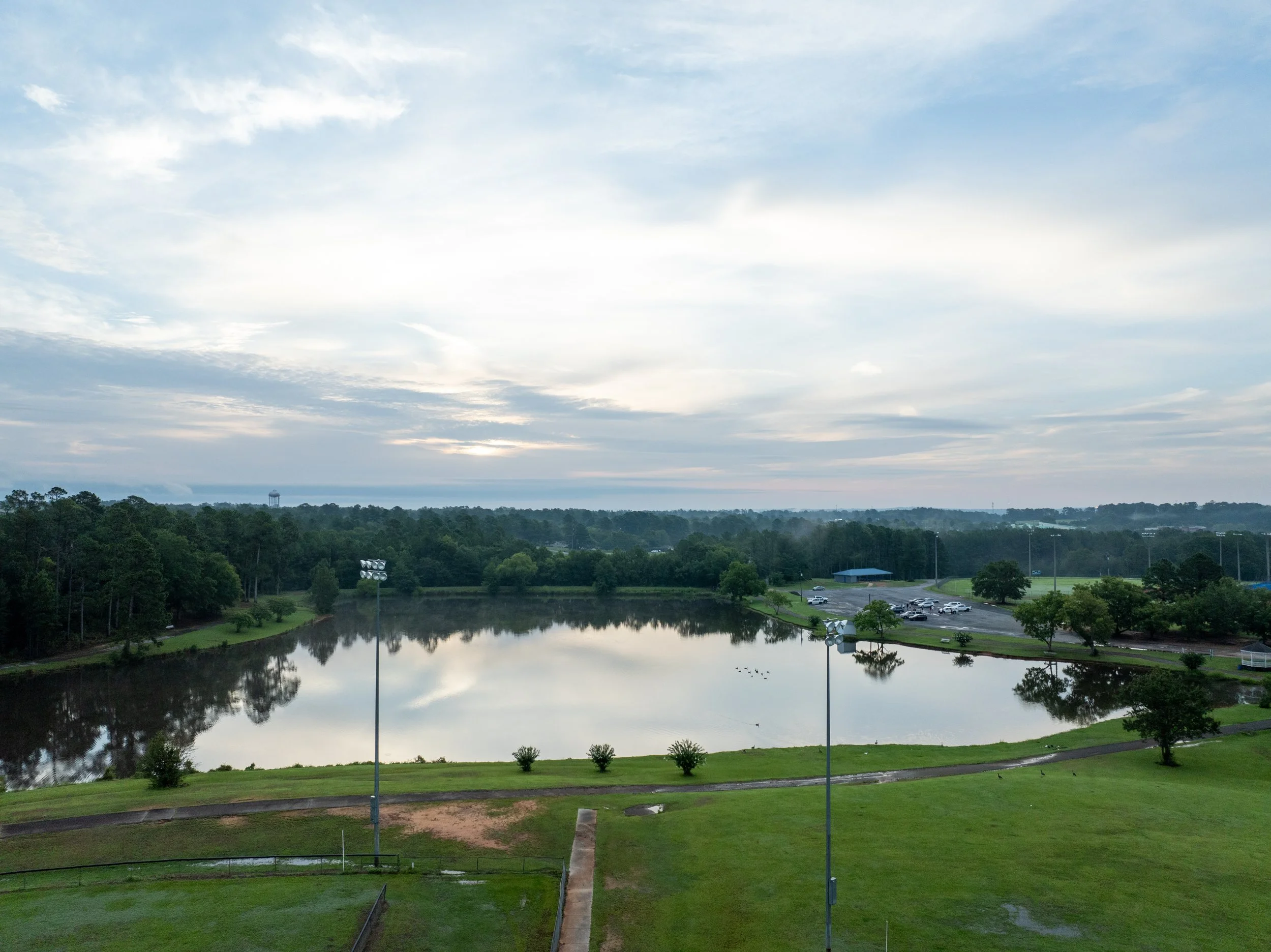 A park with a small lake, surrounded by green grass, trees, and a parking lot in the distance under a partly cloudy sky.
