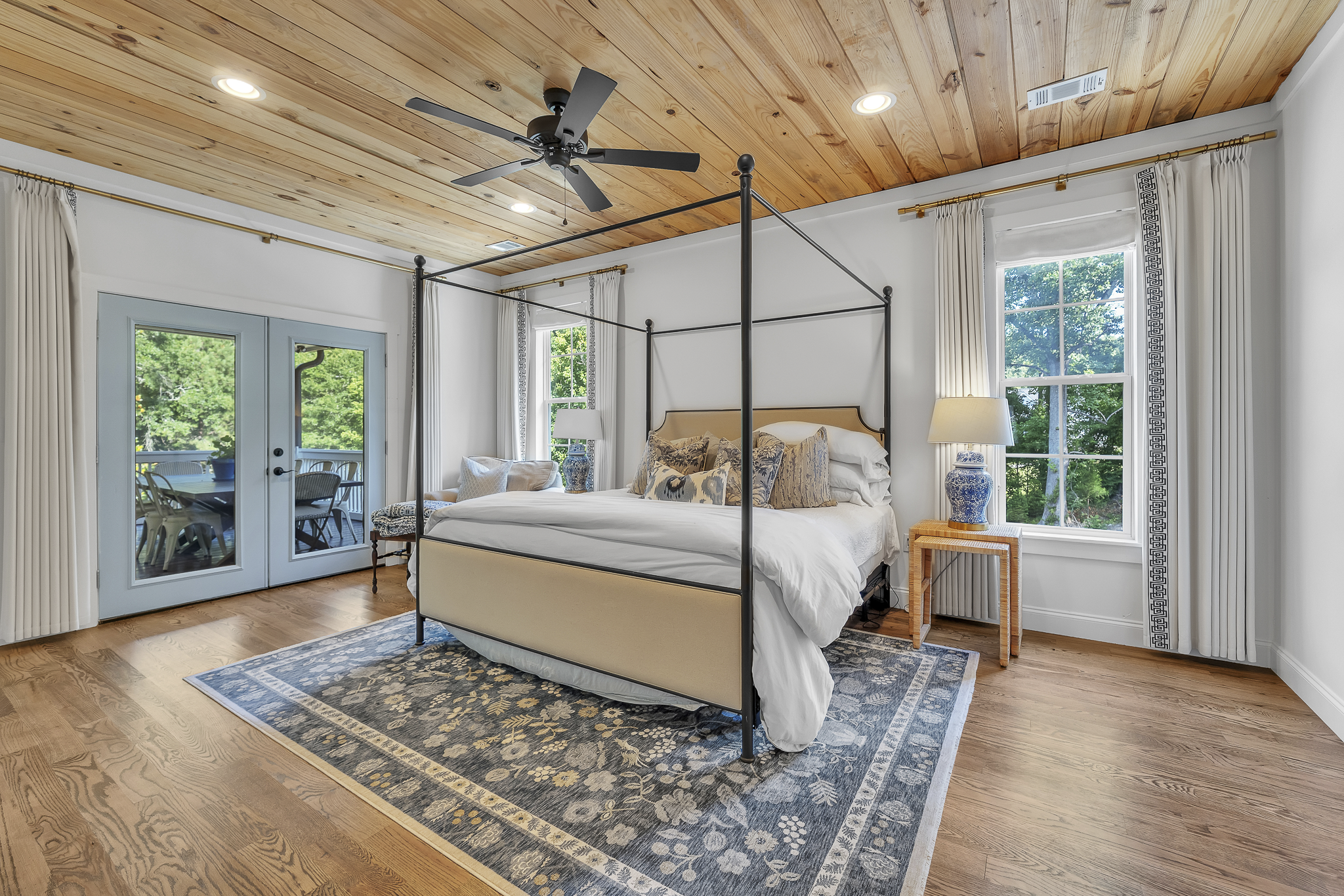 A bedroom with a four-poster bed, white bedding, decorative pillows, a wooden nightstand with a blue and white vase, and windows with curtains showing green trees outside. There is a ceiling fan and recessed lighting, with a wooden ceiling and hardwo