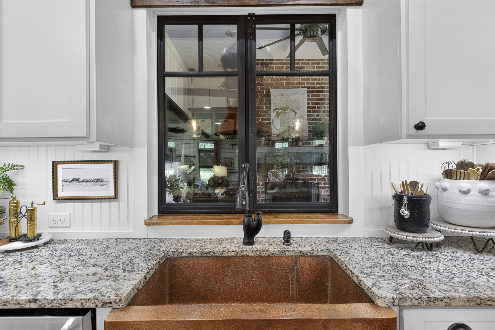 View of a kitchen window with black frame, brick wall inside, white cabinets, granite countertop, brass and white decorative items, and plants visible through the window.