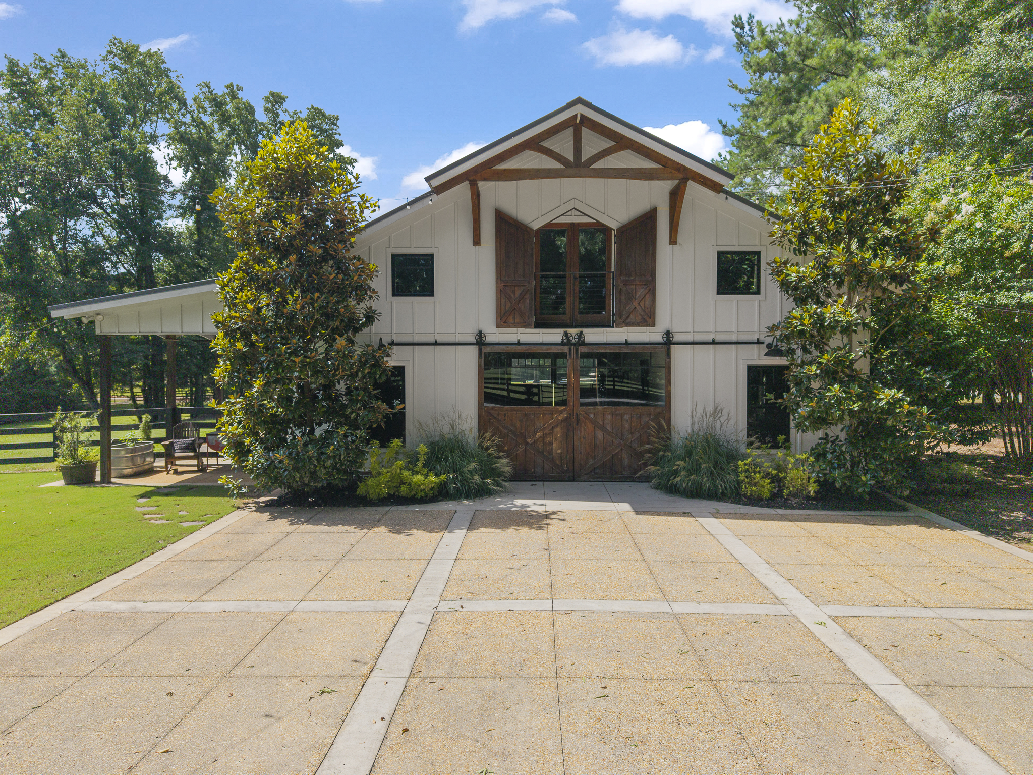A house with wood and white exterior with open barn-style doors, surrounded by green trees, a patio with outdoor furniture, and a driveway, under a partly cloudy blue sky.