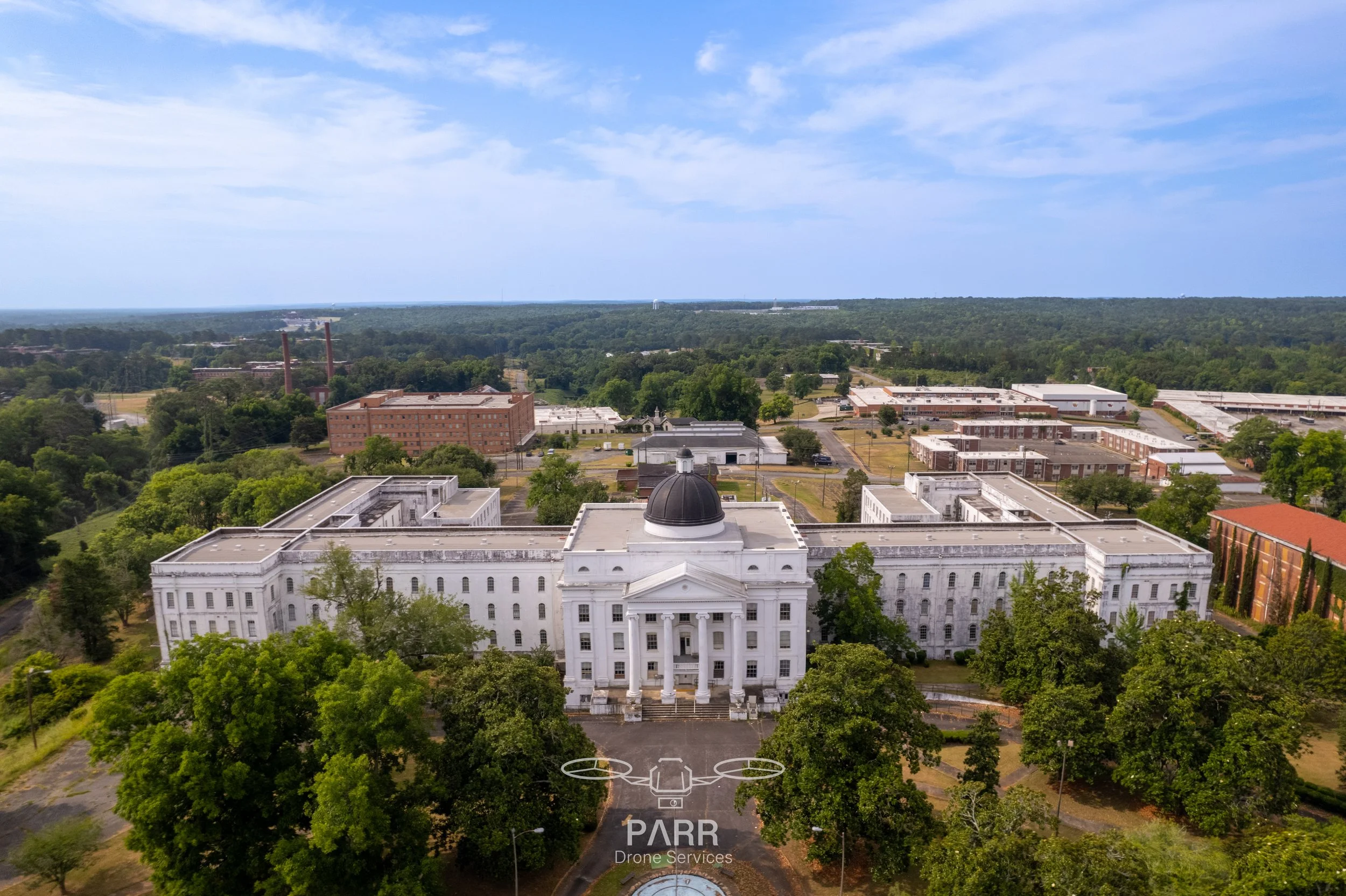 An aerial view of a historic white government building with a black dome, surrounded by green trees, with a background of a small town and forest under a partly cloudy sky.