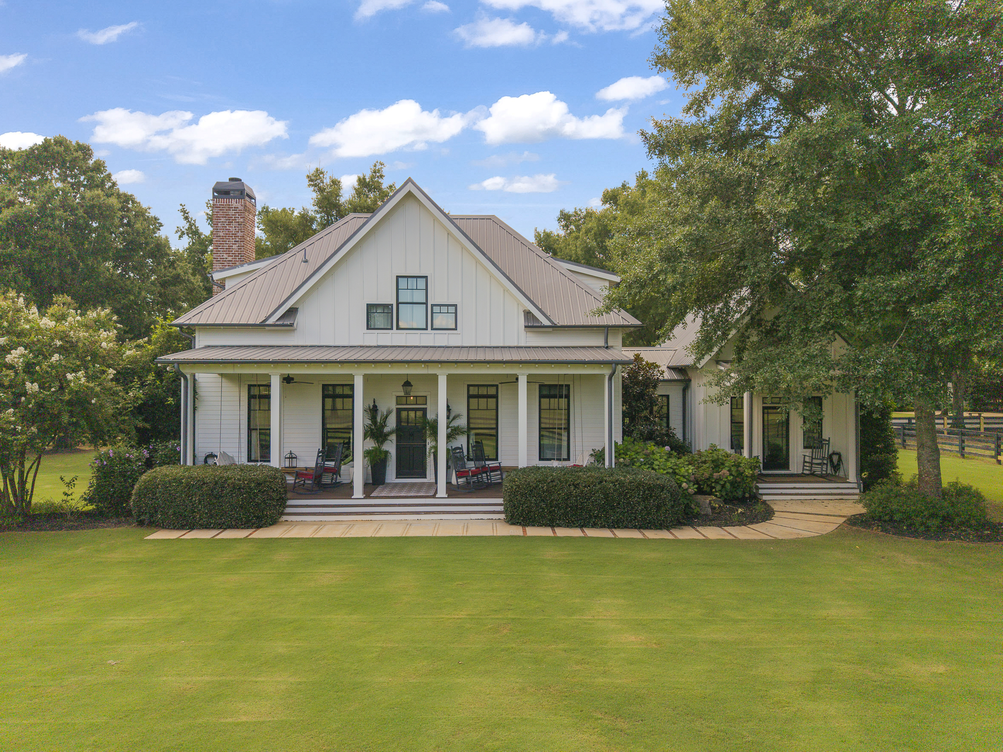 Front view of a large white house with a metal roof, porch with chairs and potted plants, surrounded by green trees and a well-maintained lawn, under a partly cloudy sky.