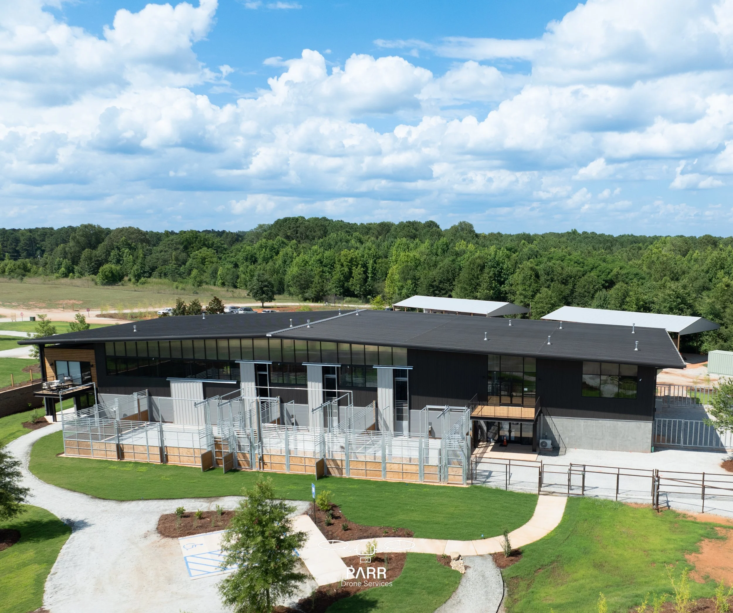 Modern building with large glass windows, black exterior, surrounded by landscaped stairs, pathways, and green lawns, with trees and a blue sky with clouds in the background.