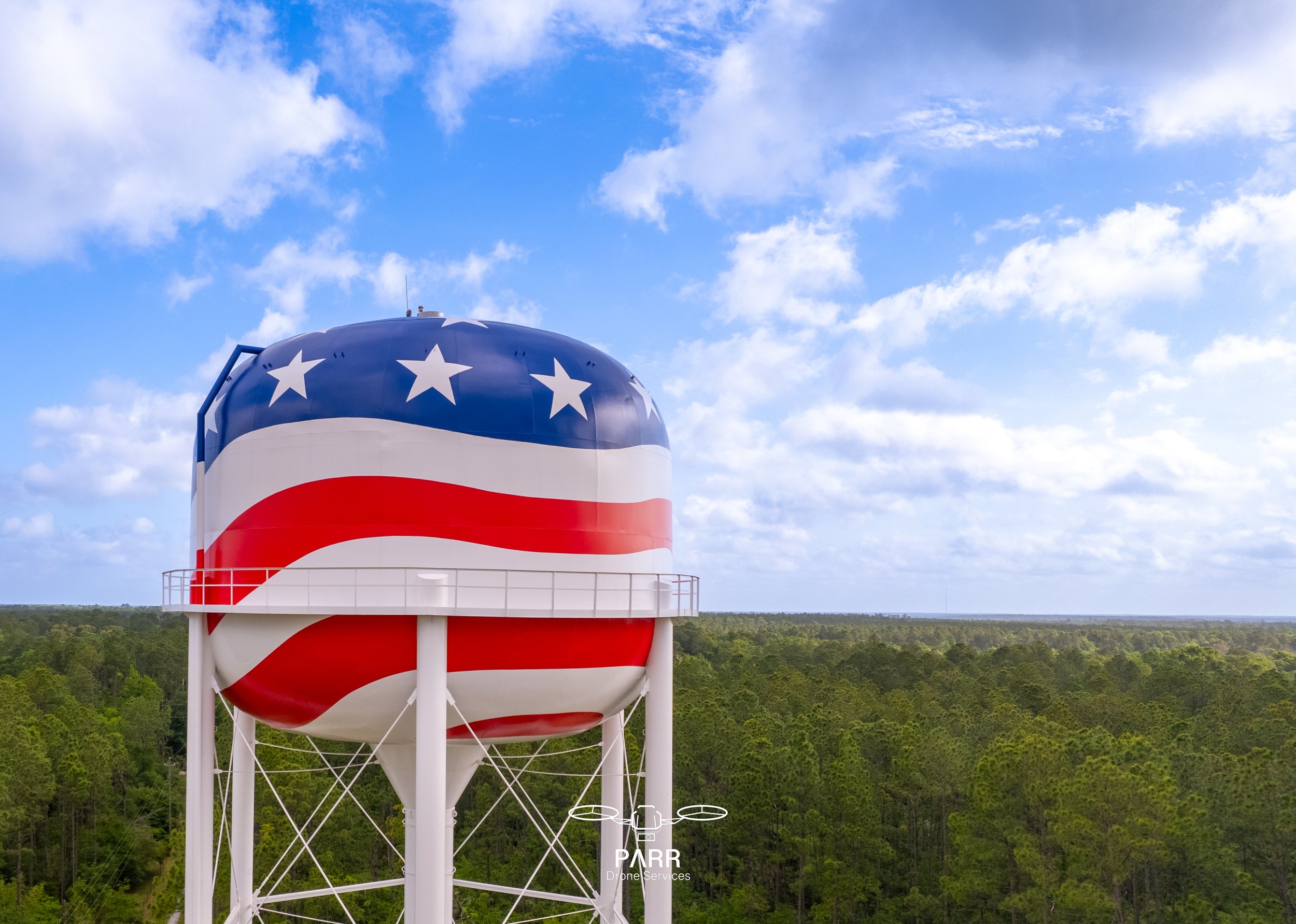 Water tower painted with American flag design, featuring stars and stripes, standing above a green forest with a blue sky and scattered clouds in the background.