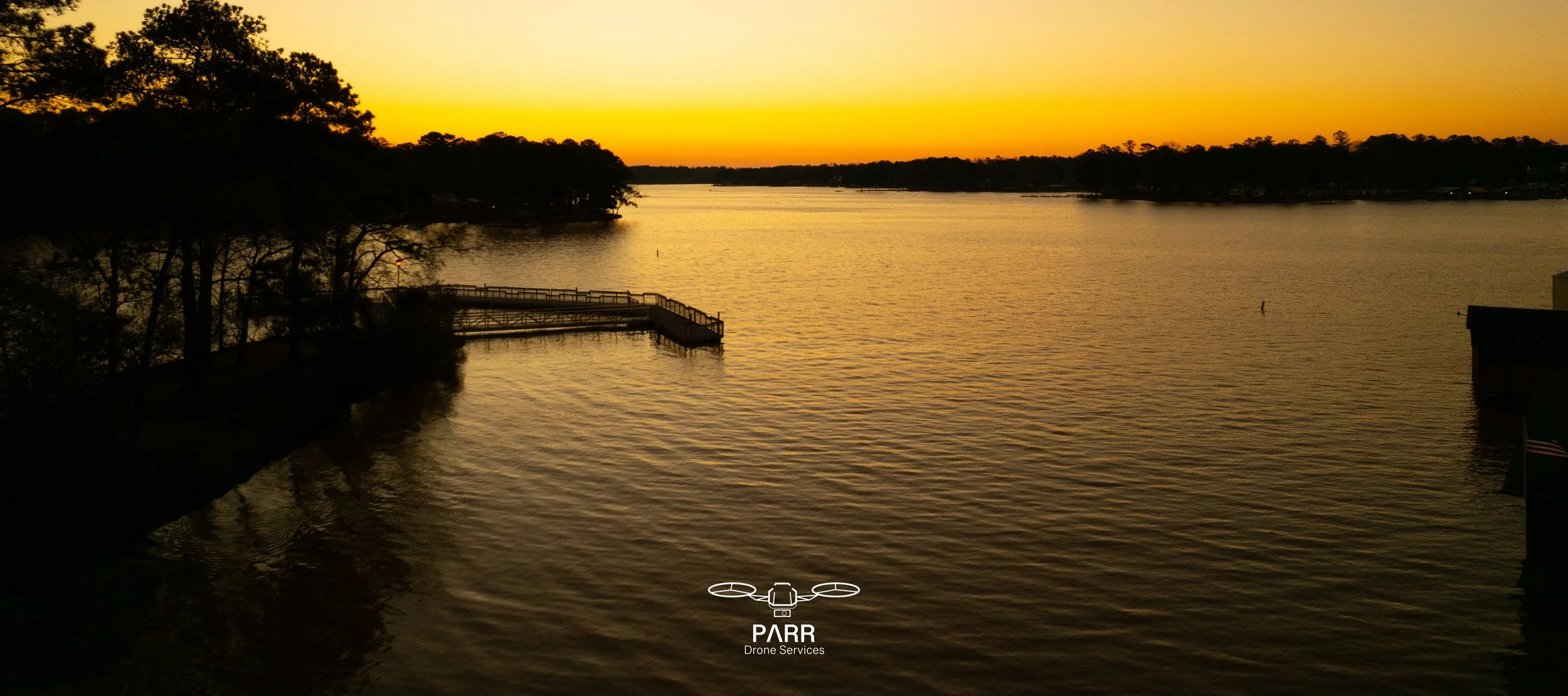 Sunset over a body of water with trees on the left and right, and a dock extending into the water in the foreground. The sky is orange and yellow, reflecting on the rippling water.